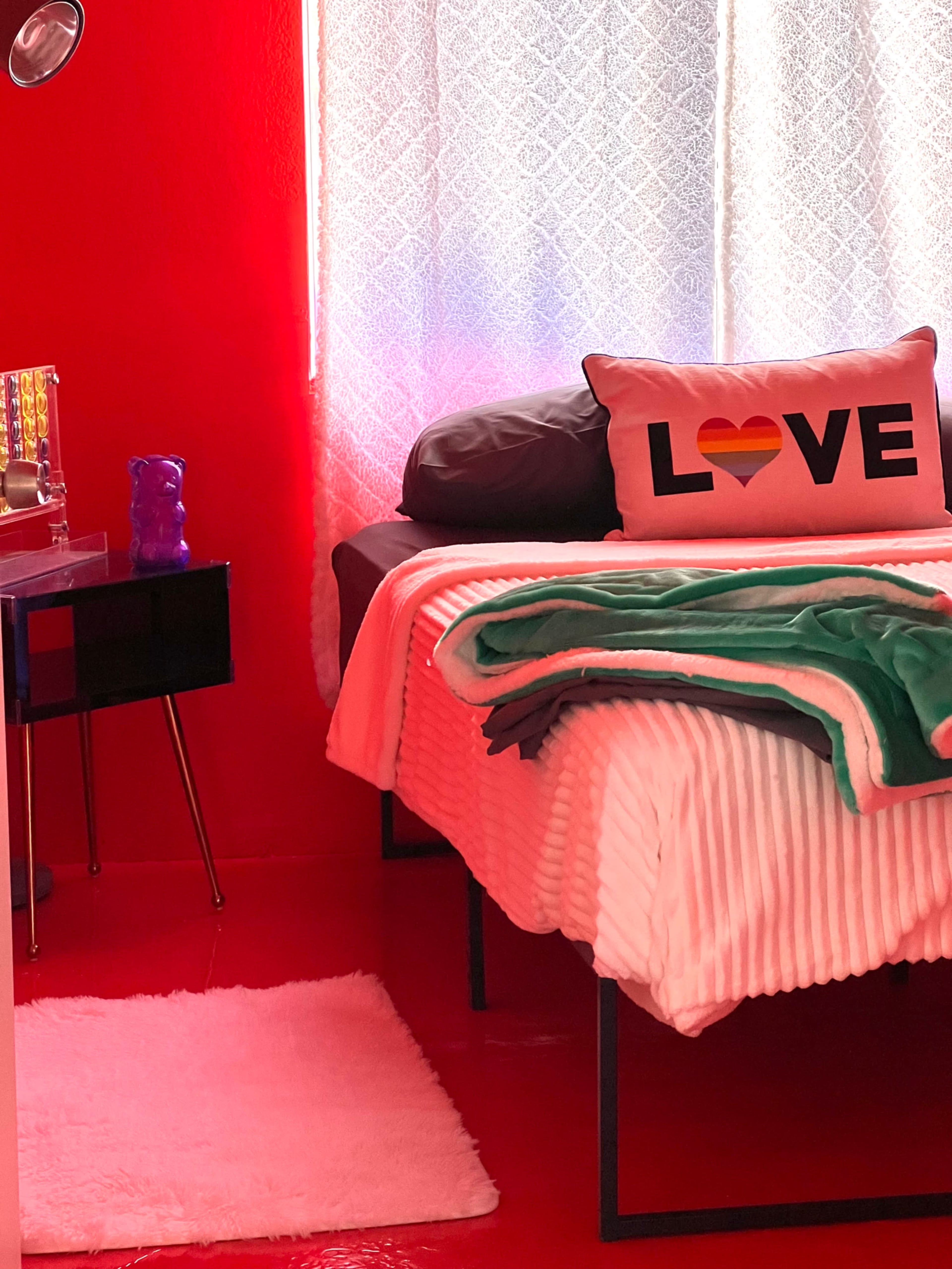 A bedroom with a red wall, a bed covered with a white striped blanket, and a "LOVE" decorative pillow on top.