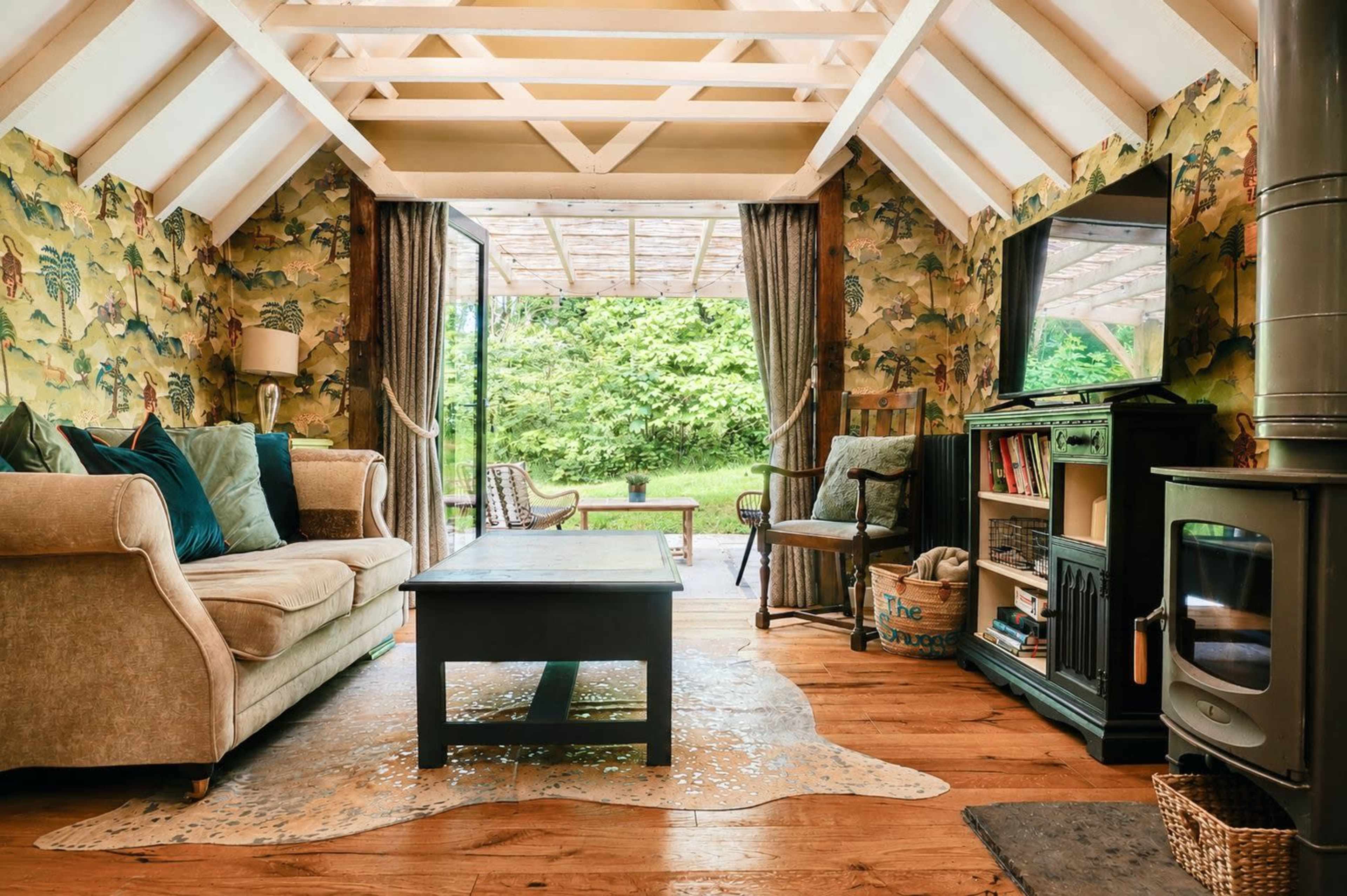 The image shows a cozy living room with a floral wallpaper, a sofa, a wooden coffee table, and large windows that open to a green outdoor space.