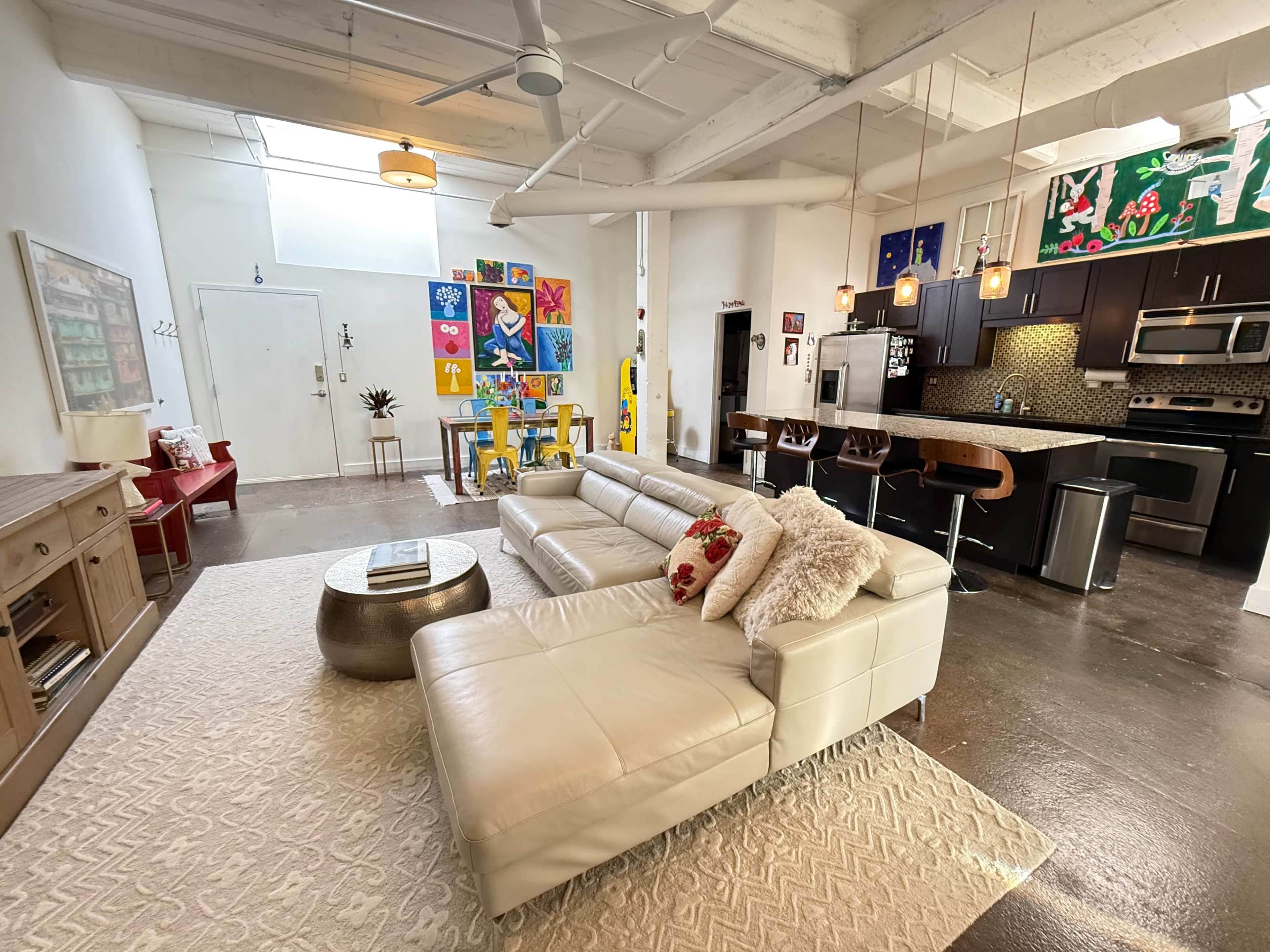 The image shows a modern loft-style living area with a sectional sofa, a decorative coffee table, and an open kitchen featuring bar seating and colorful artwork on the walls.