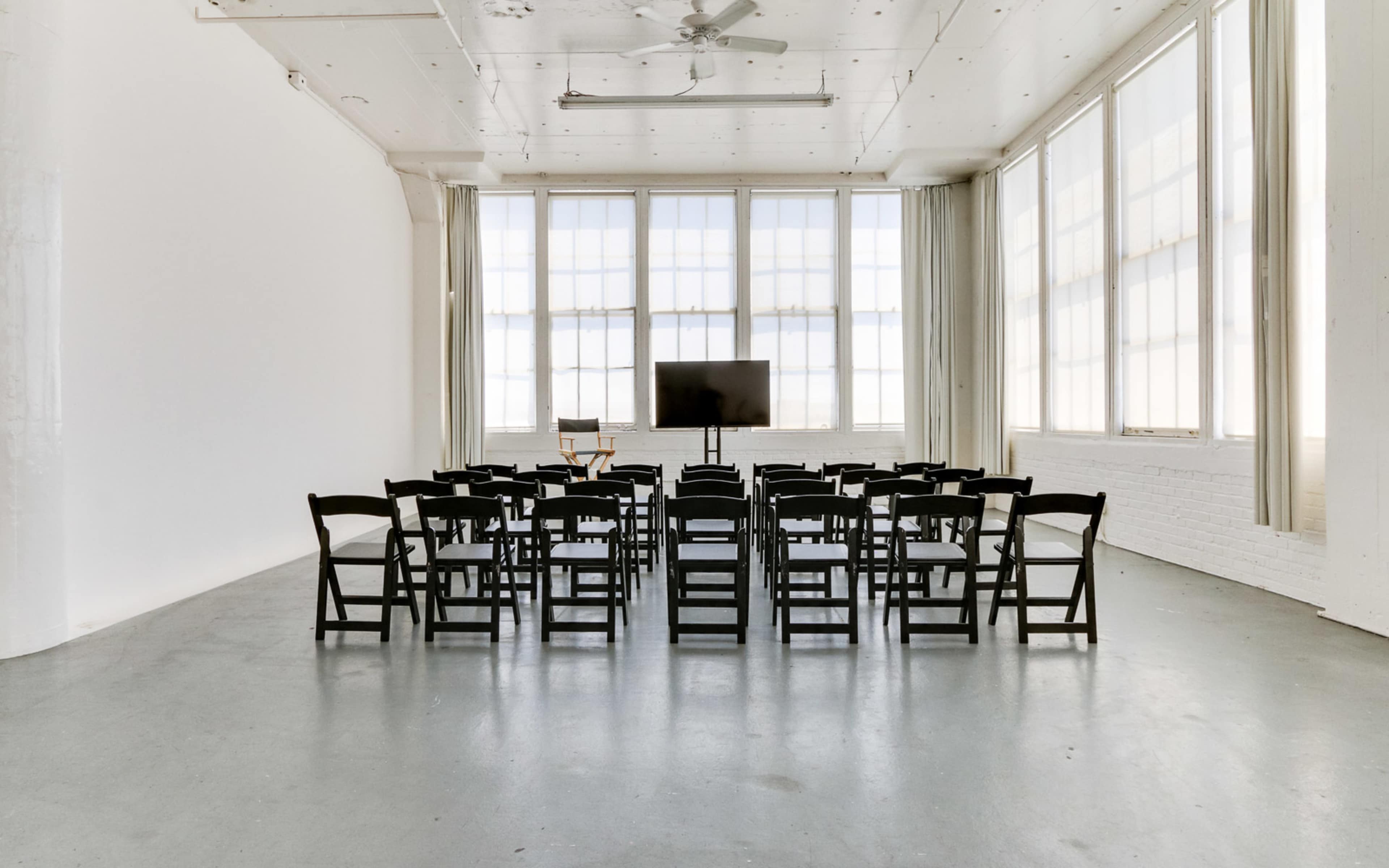 A minimalist meeting room features rows of black chairs arranged around a monitor in front of large windows.