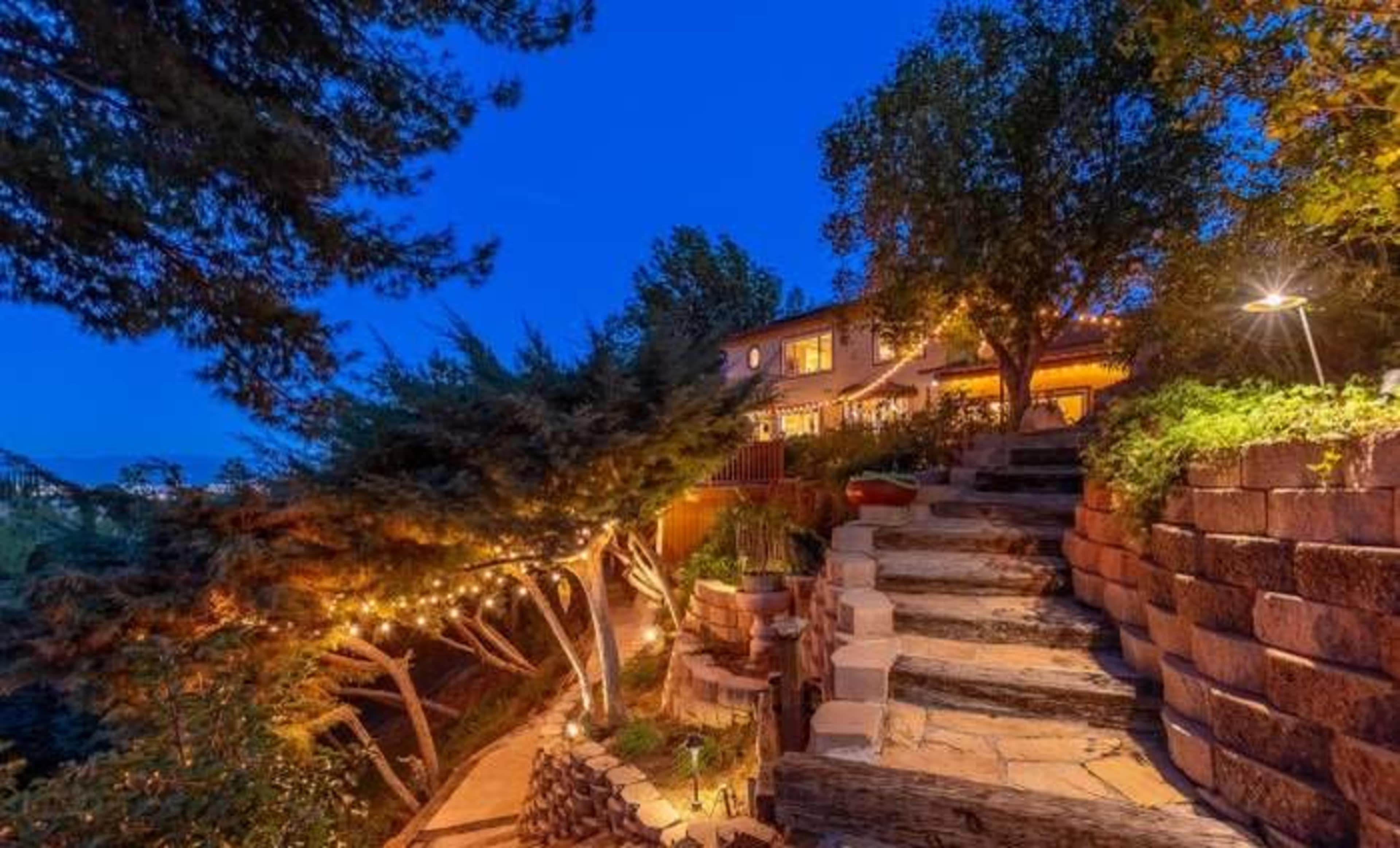 A winding stone pathway leads up to a house nestled among trees, illuminated by warm lights against a twilight backdrop.