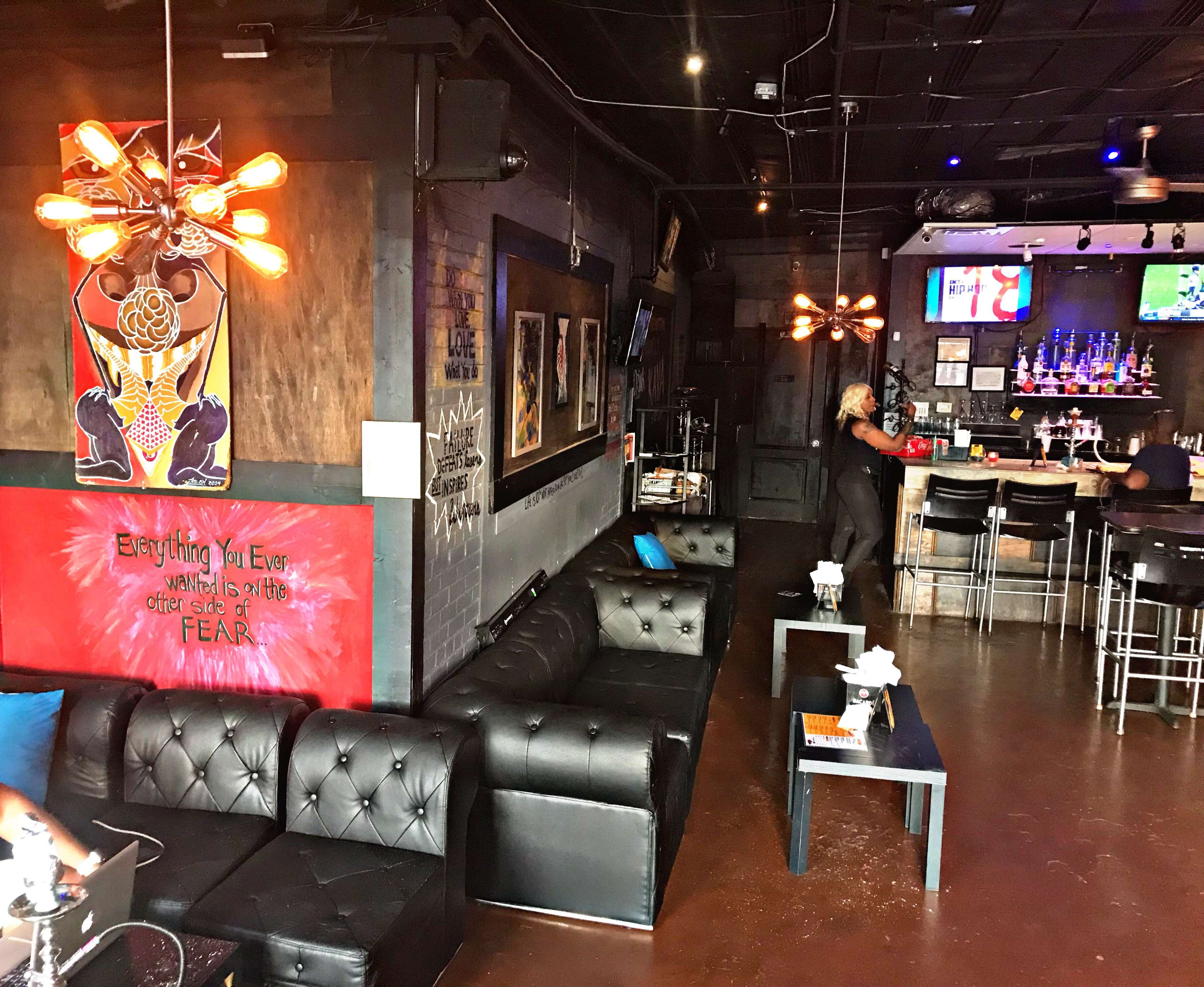 A cozy lounge features black leather couches, a bar area with high stools, and eclectic wall art illuminated by industrial-style light fixtures.
