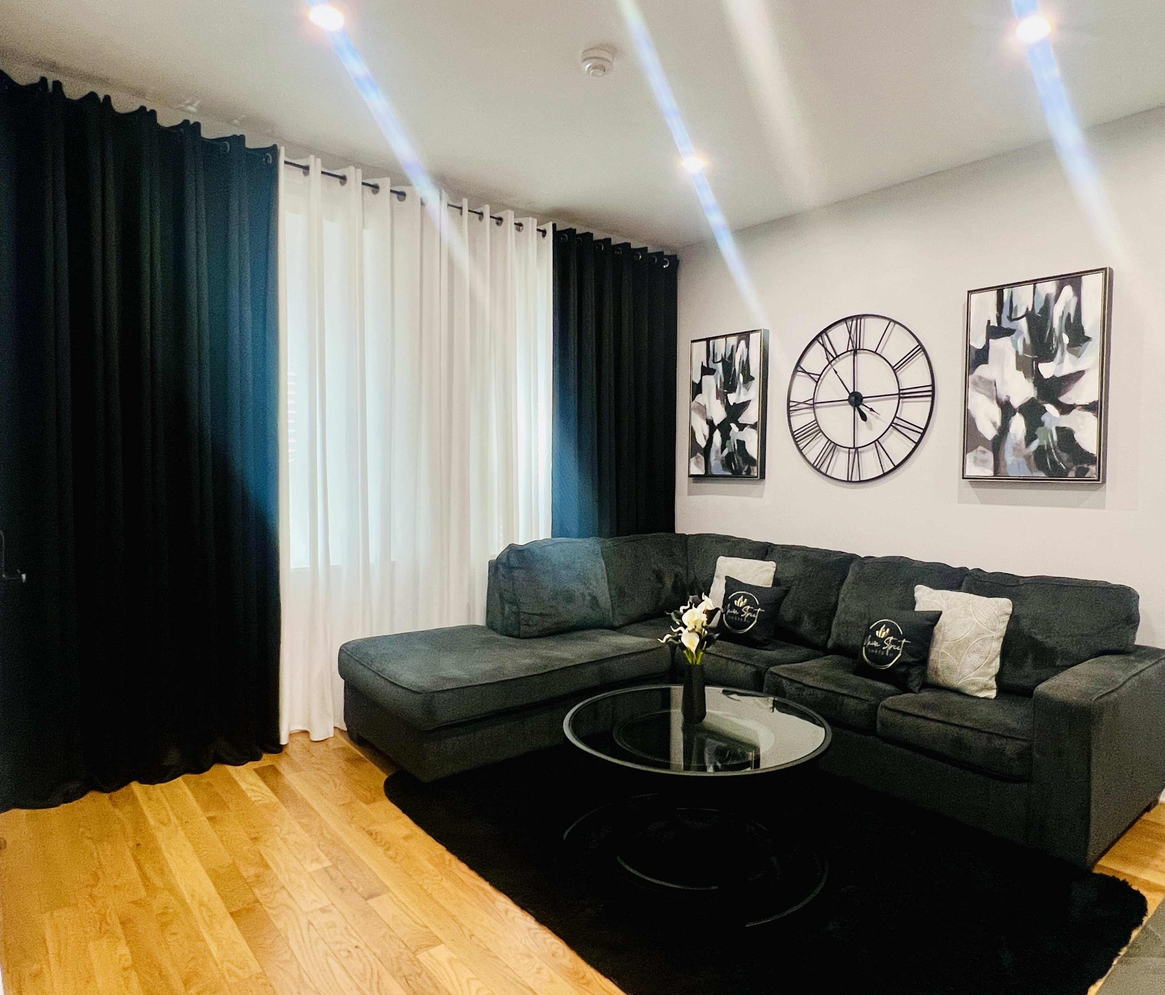 The image shows a modern living room featuring a dark sectional sofa, a round glass coffee table, and black and white curtains, with artwork and a large wall clock hanging on the walls.