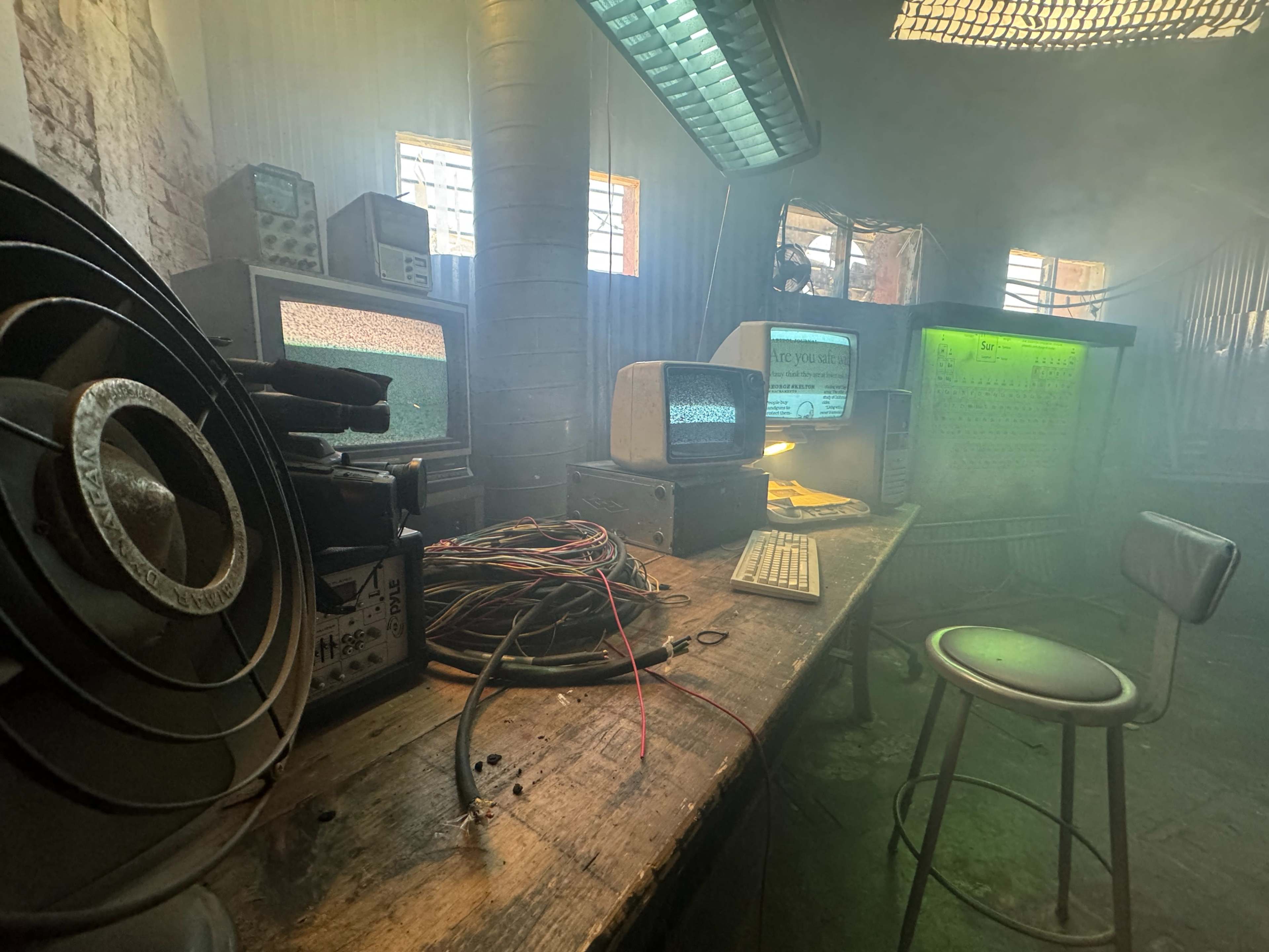 The image shows a dimly lit room with an old wooden table covered in computer equipment, wires, and a chair, alongside multiple vintage monitors displaying static.