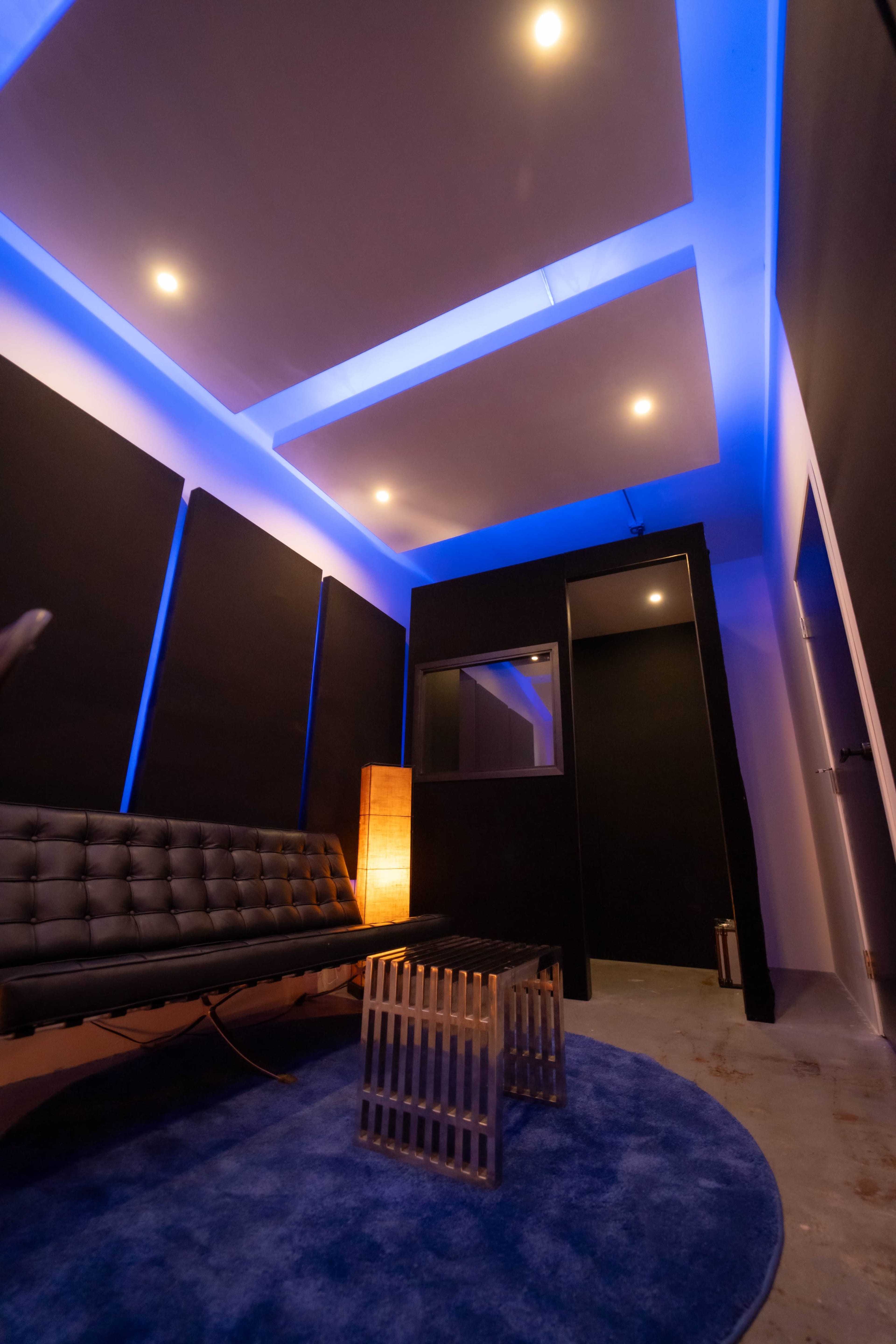A modern room features a black couch, a wooden side table, blue ambient lighting, and black acoustic wall panels.