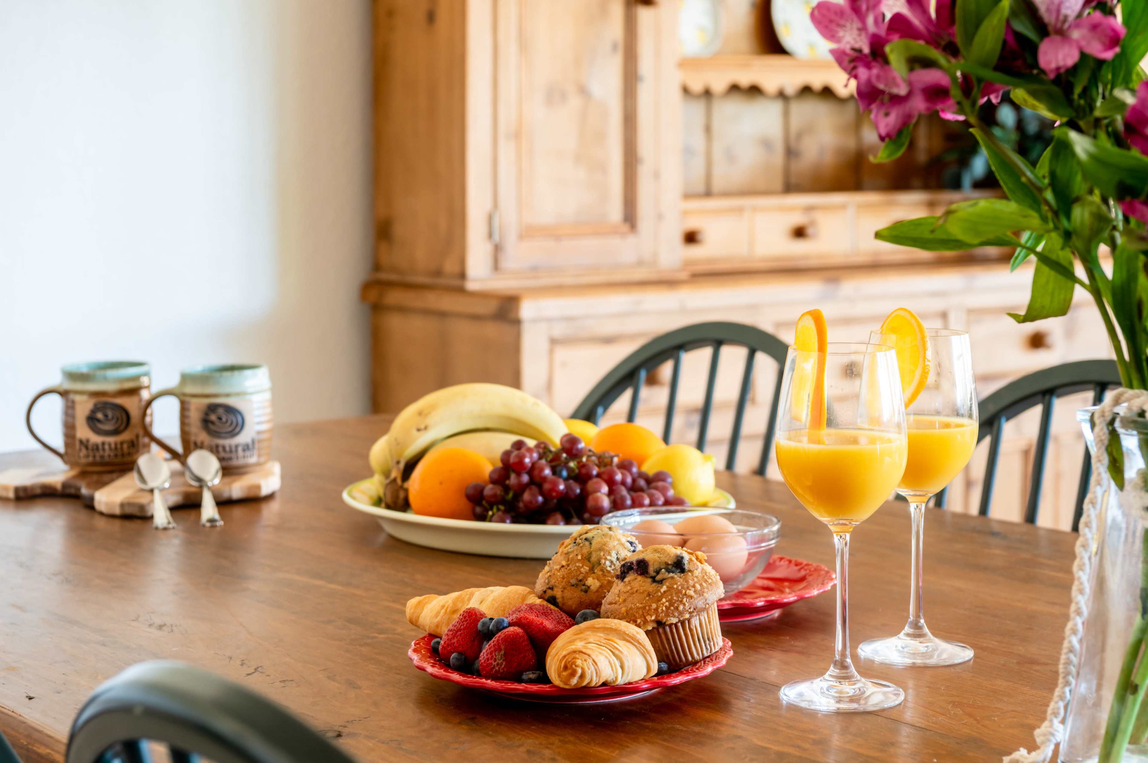 A wooden dining table is set with a plate of muffins and pastries, two glasses of orange juice garnished with orange slices, and a bowl of fresh fruit.