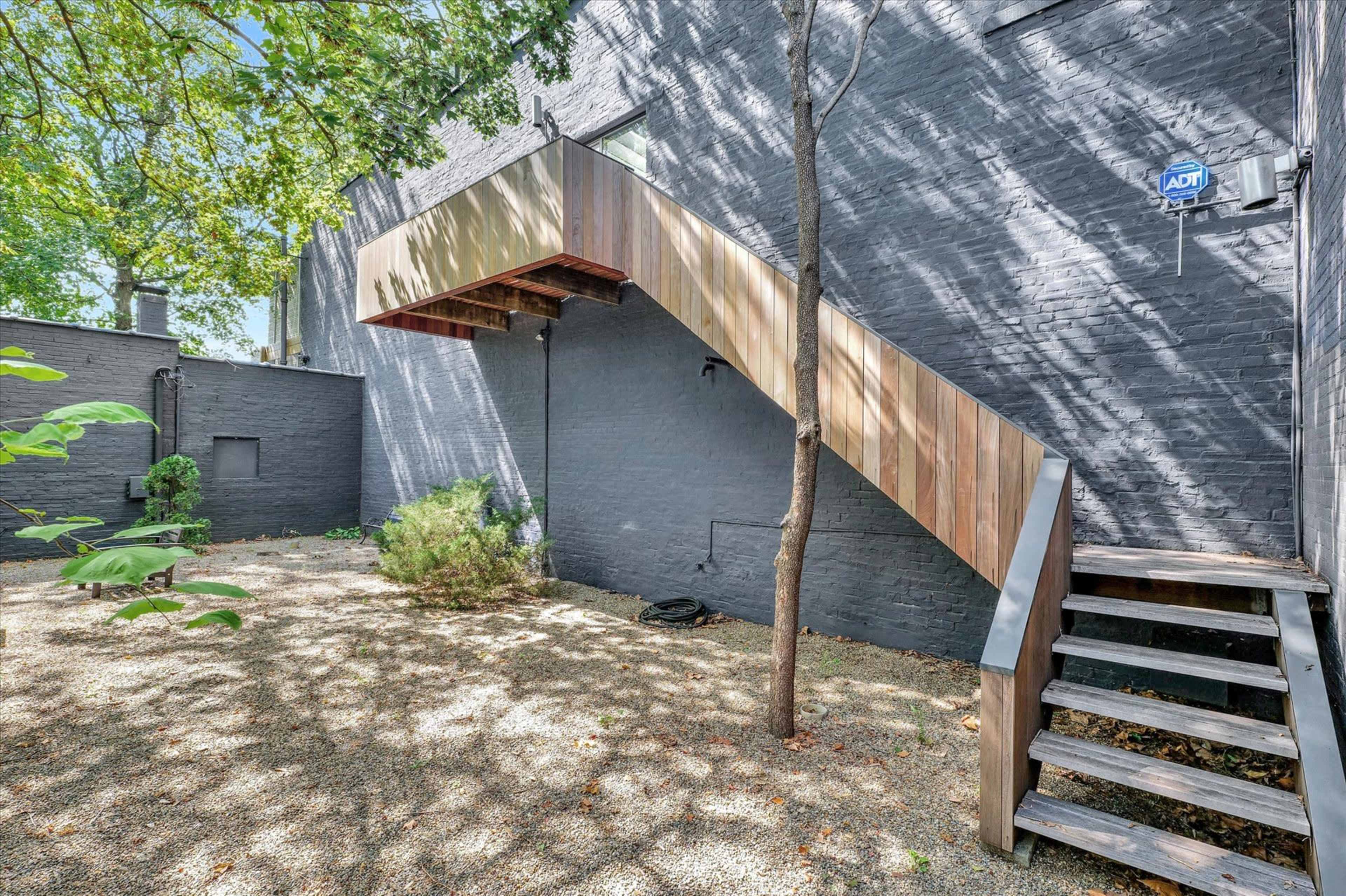 A wooden stairway leading to an upper entrance, beside a stone wall in a garden area with gravel and sparse greenery.