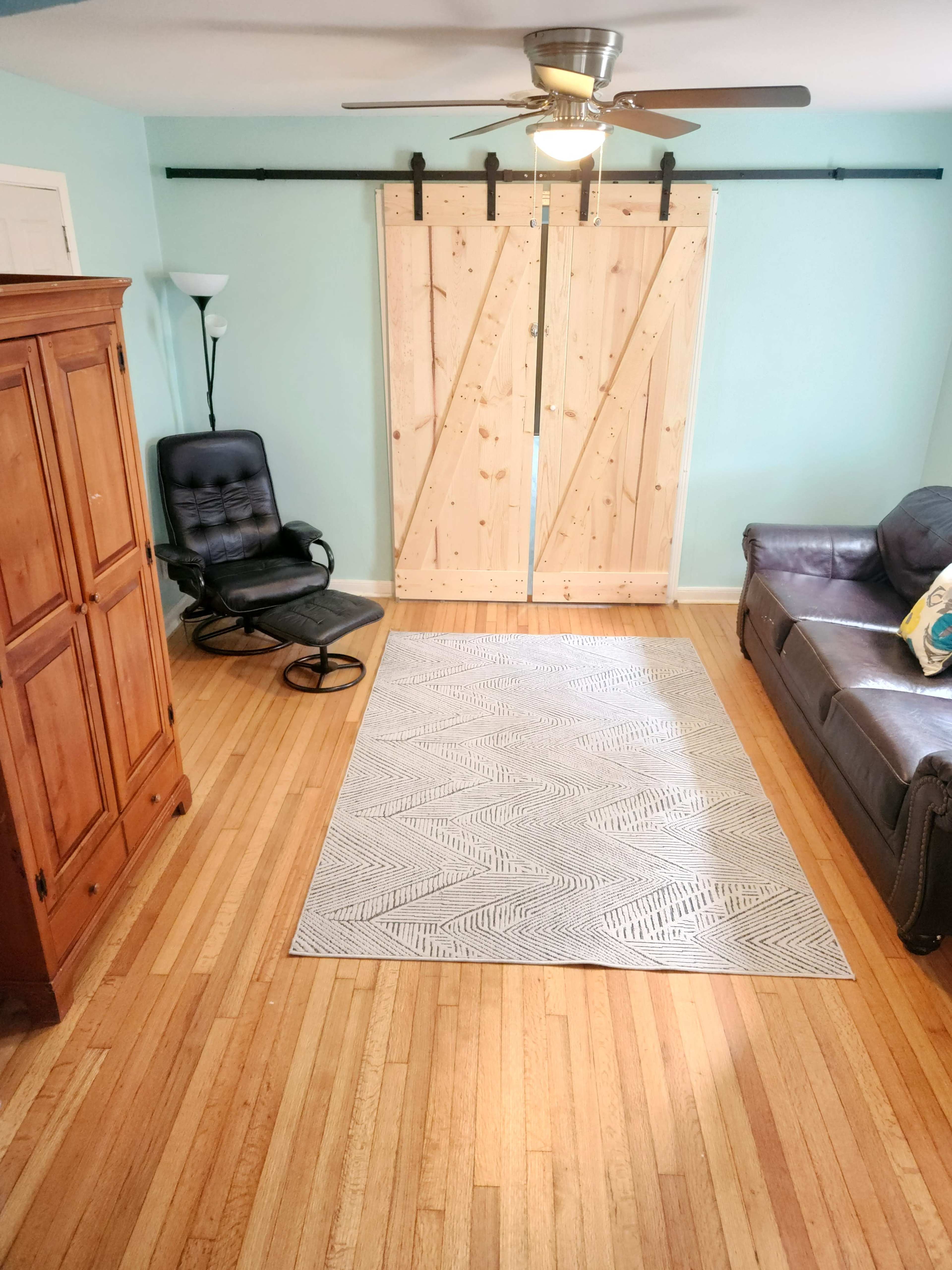 The room features a leather sofa, a black rocking chair, a wooden wardrobe, a patterned area rug, and sliding barn doors against a light blue wall.
