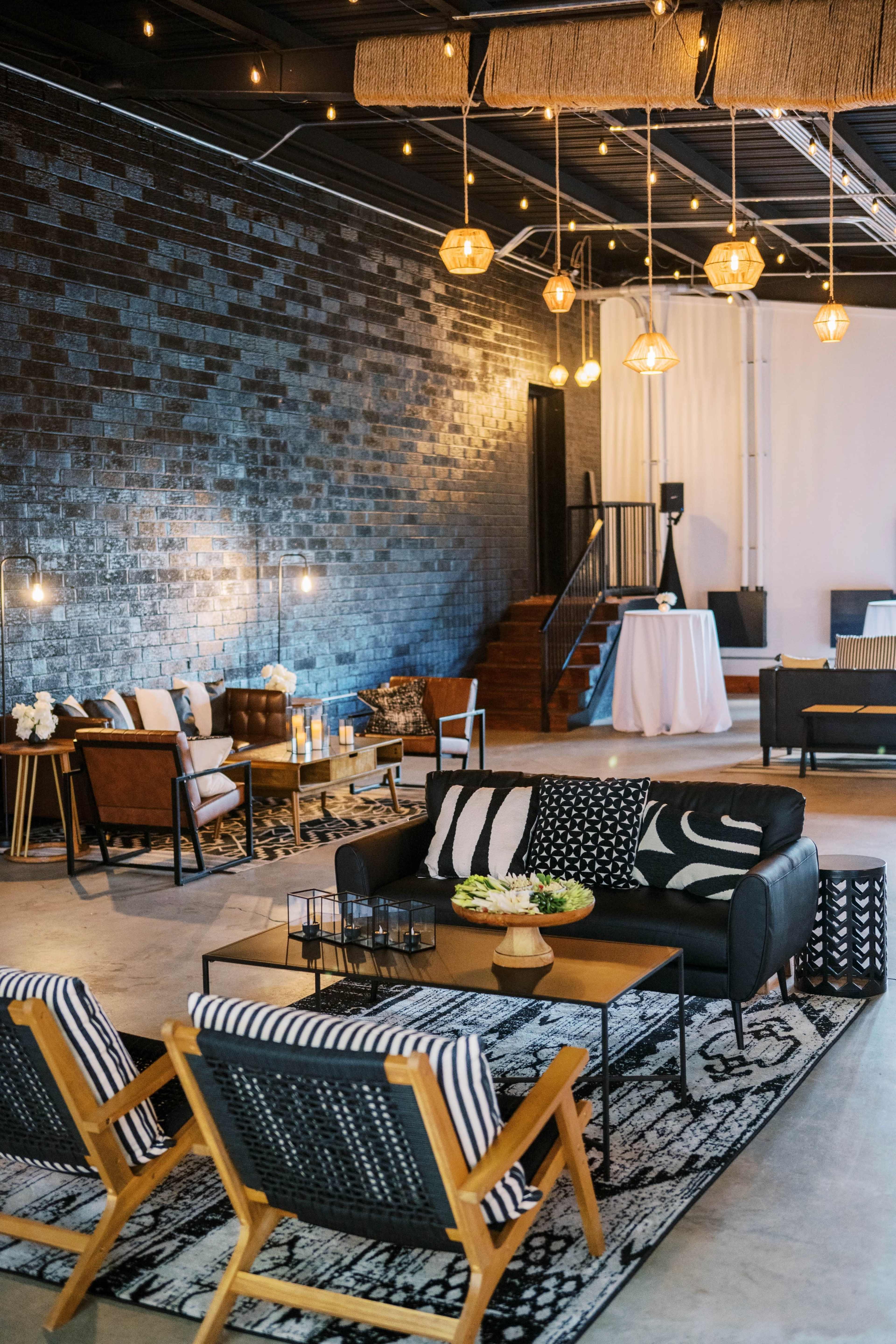 The image shows a modern lounge area with a combination of black and wooden furniture, decorative lighting, and a patterned rug on a concrete floor.