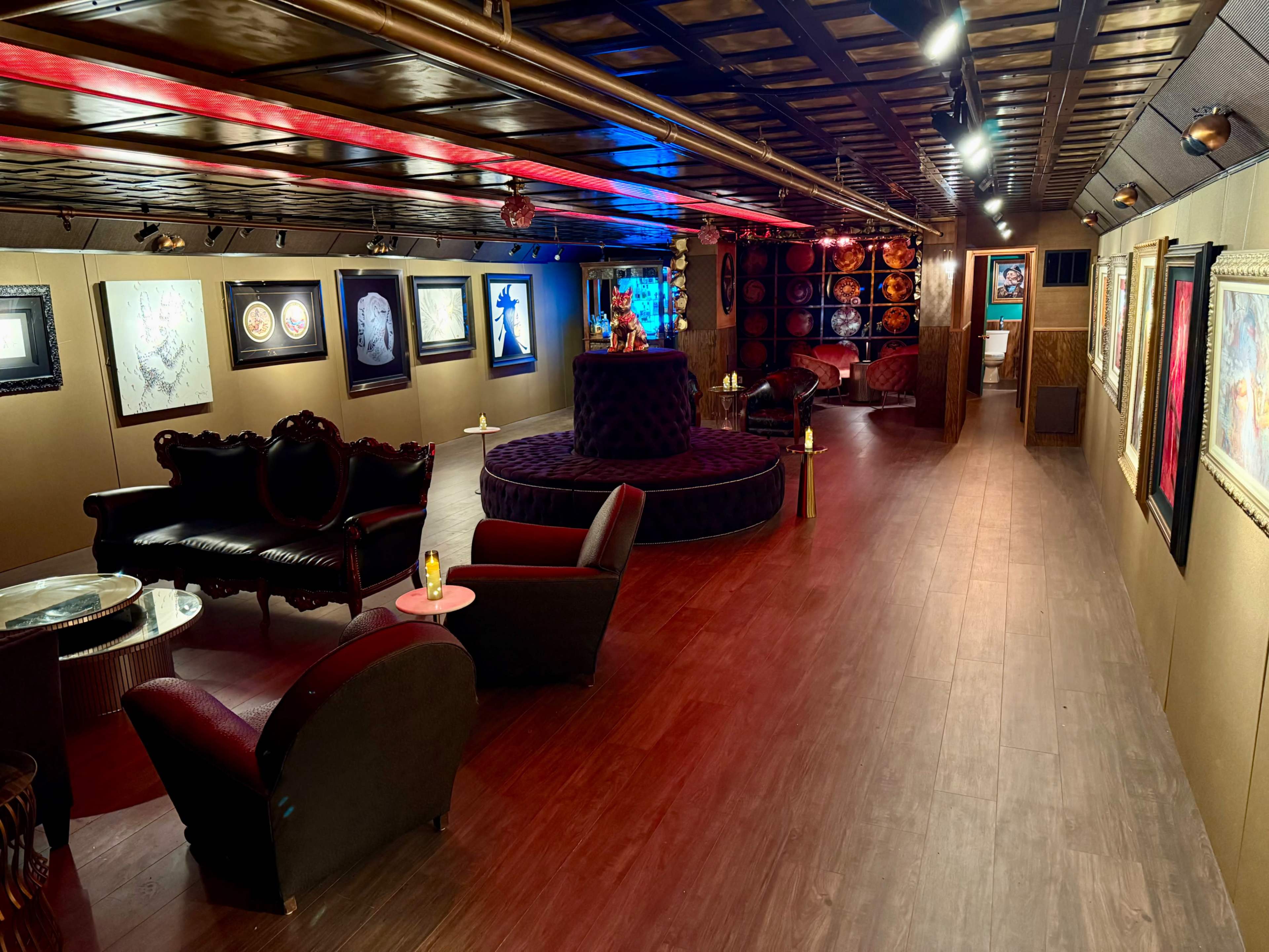 The image shows a stylishly designed lounge area featuring dark furnishings, artwork on the walls, and a central circular seating arrangement.