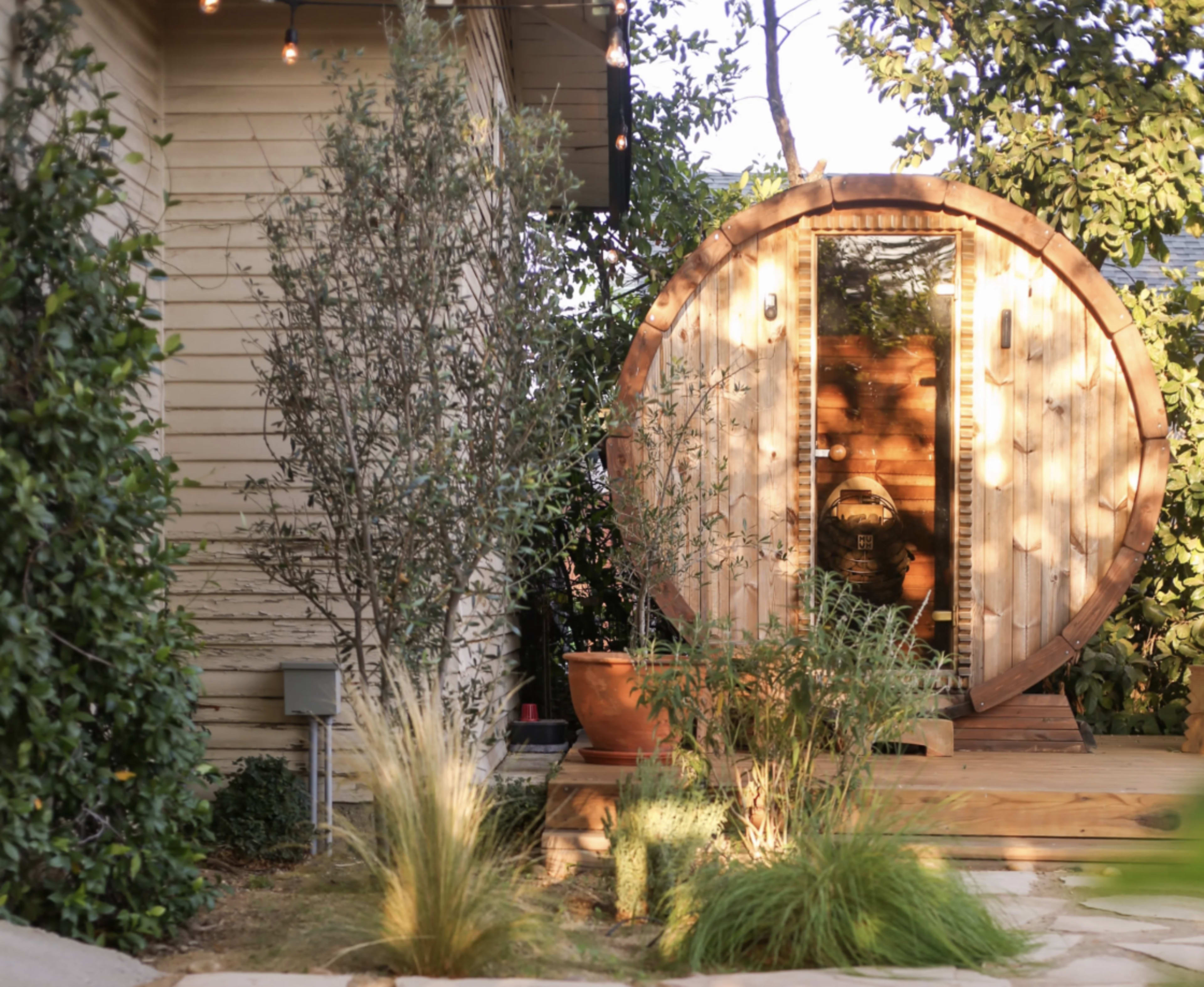 The image features a circular wooden sauna structure situated next to a house, surrounded by various plants and decorative lighting.