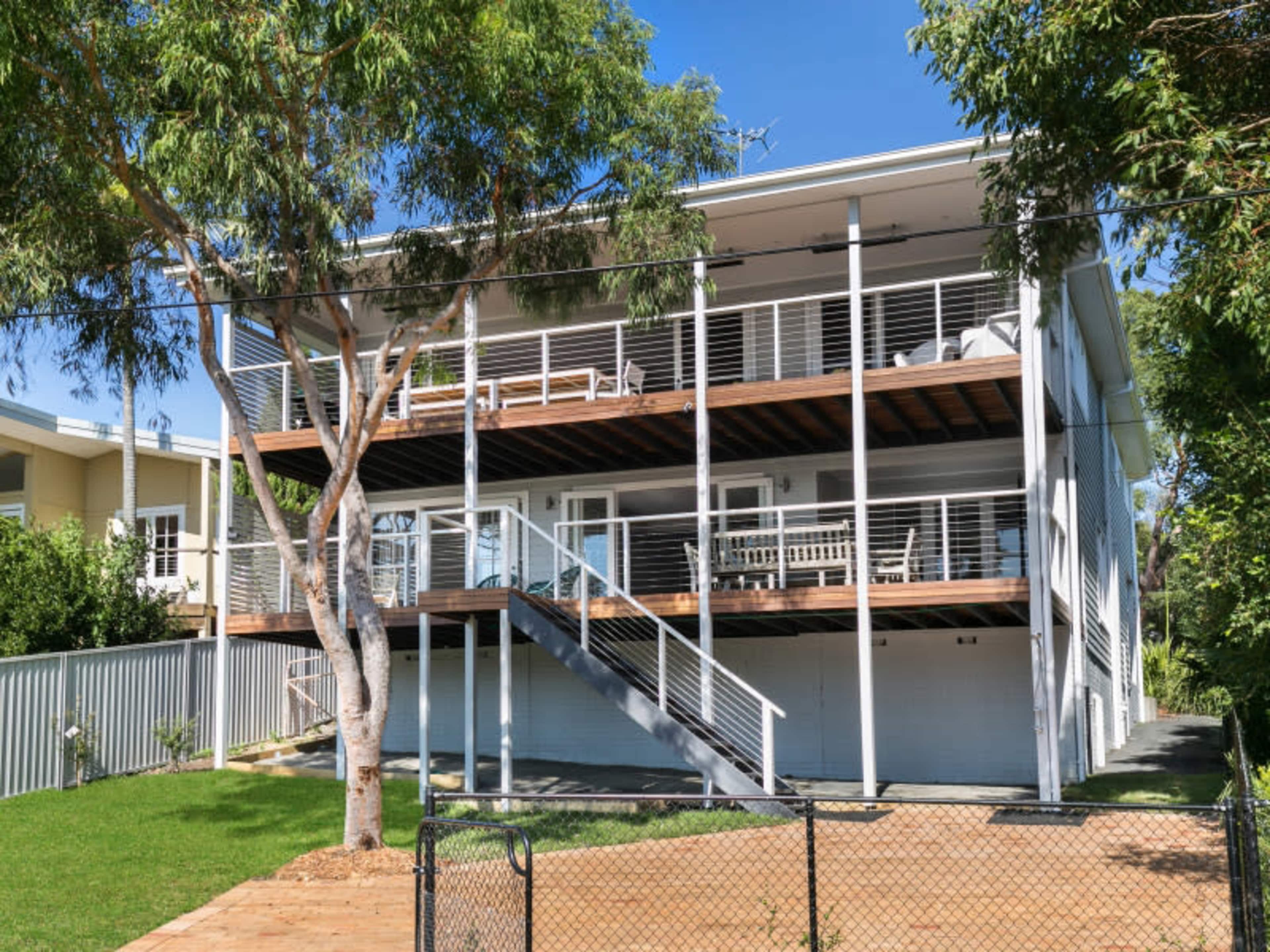 A three-story house with balconies and a wooden deck is set on a landscaped yard featuring a tree and a fenced pathway.