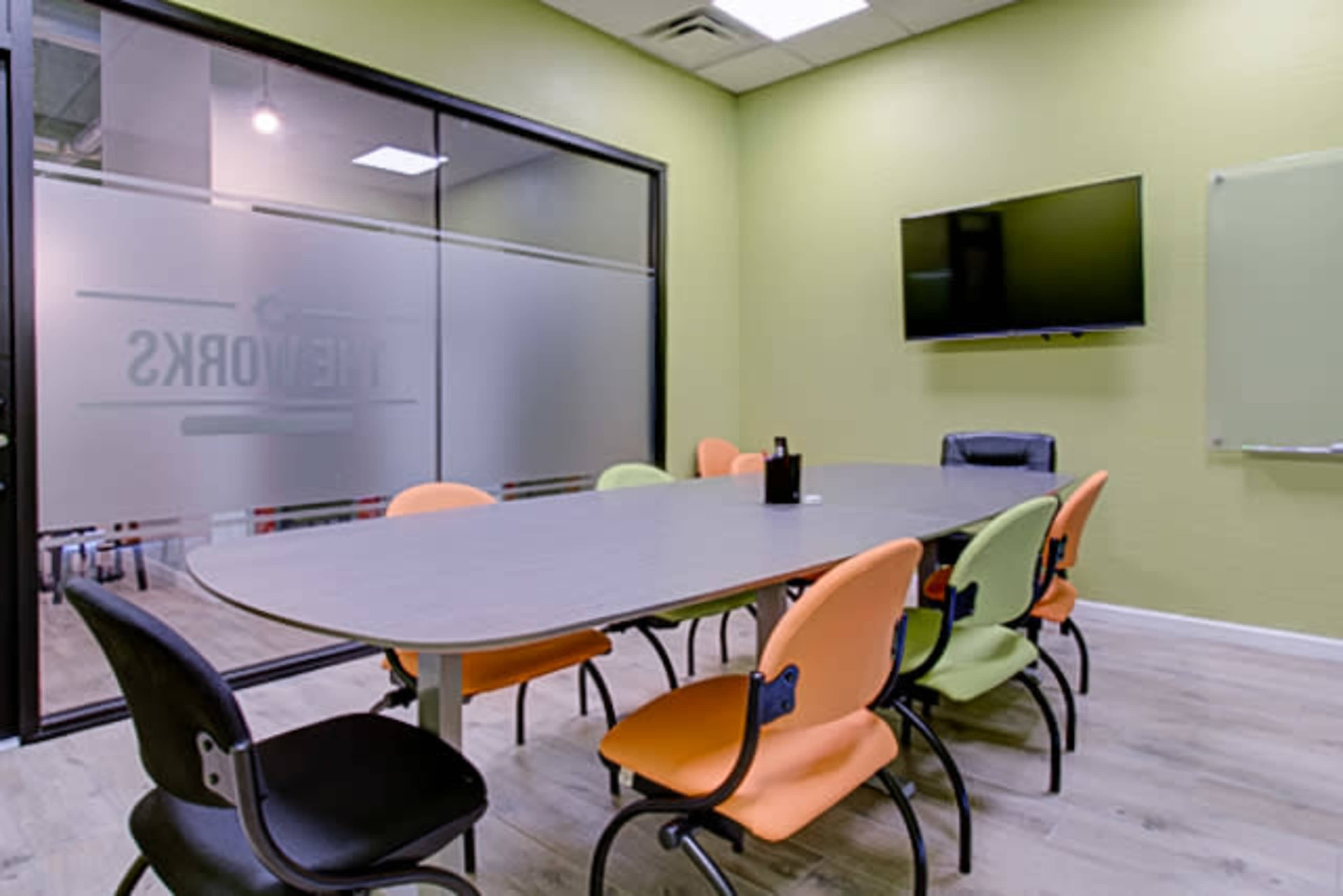 A meeting room features a large table surrounded by six chairs in various colors, with a television mounted on the wall and a glass partition showing the room's name.