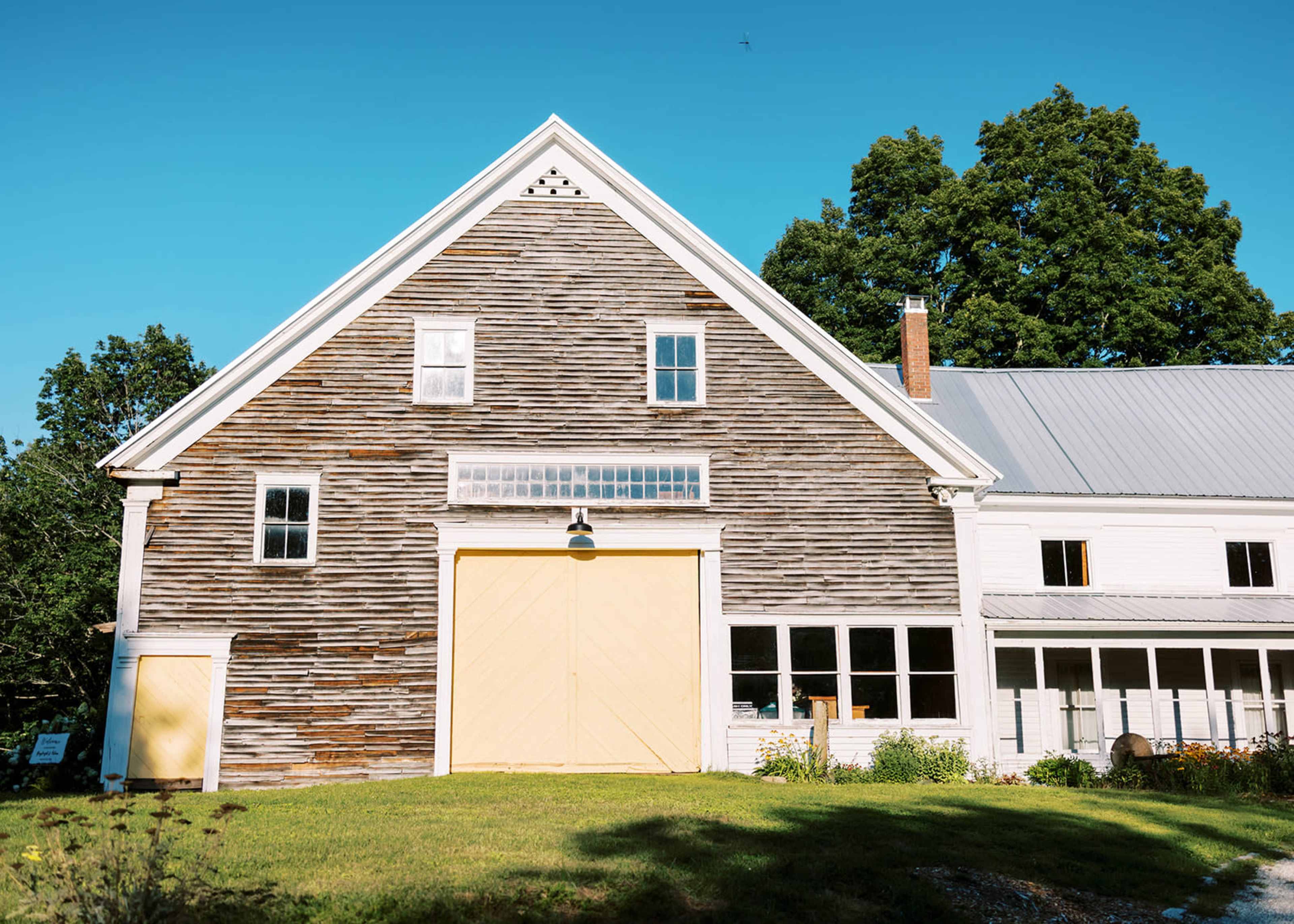 A weathered wooden barn with a yellow double door stands beside a white farmhouse under a clear blue sky.