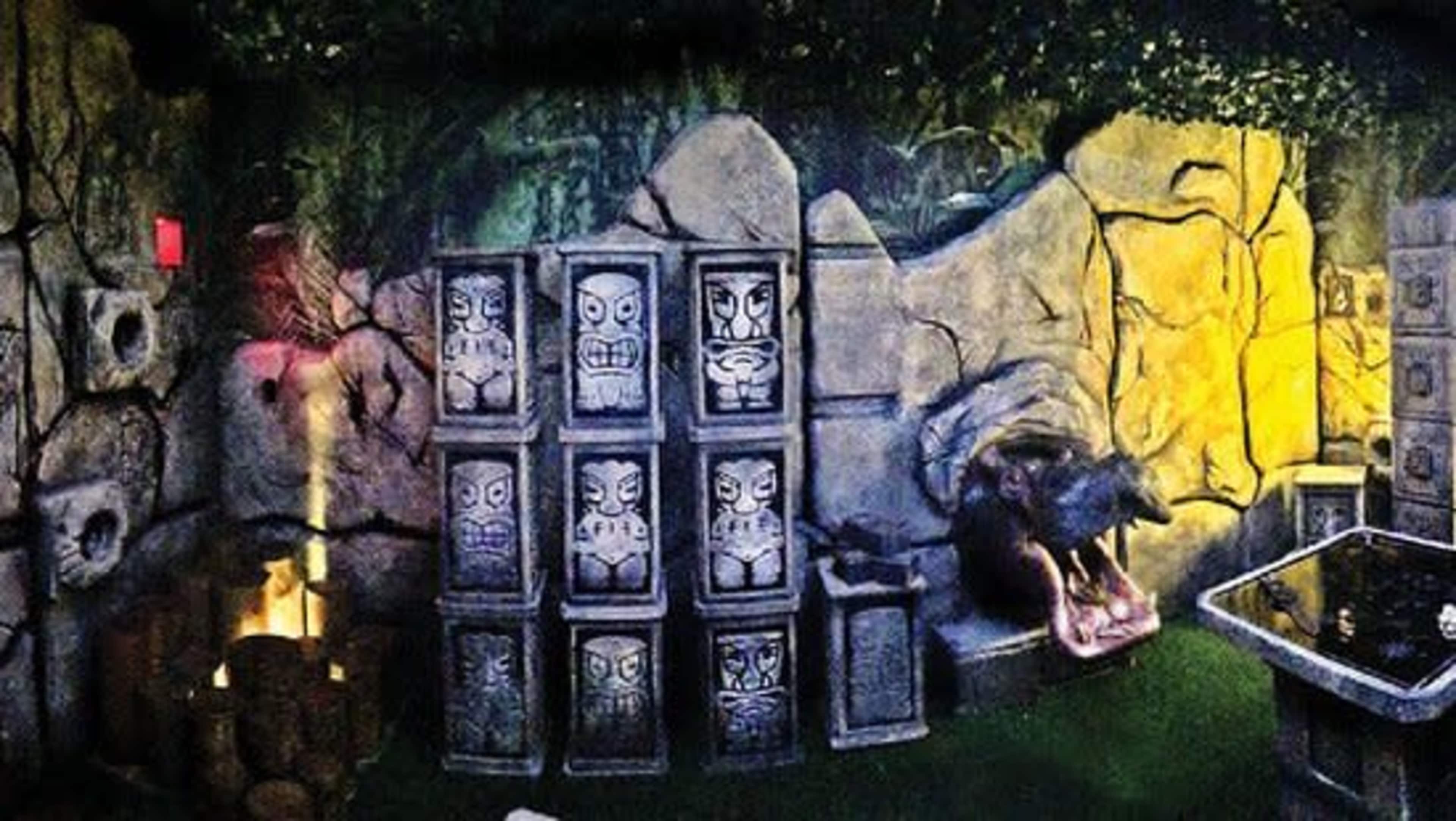 An escape room with stone walls, tiki statues, and various artifacts amid a dimly lit setting.
