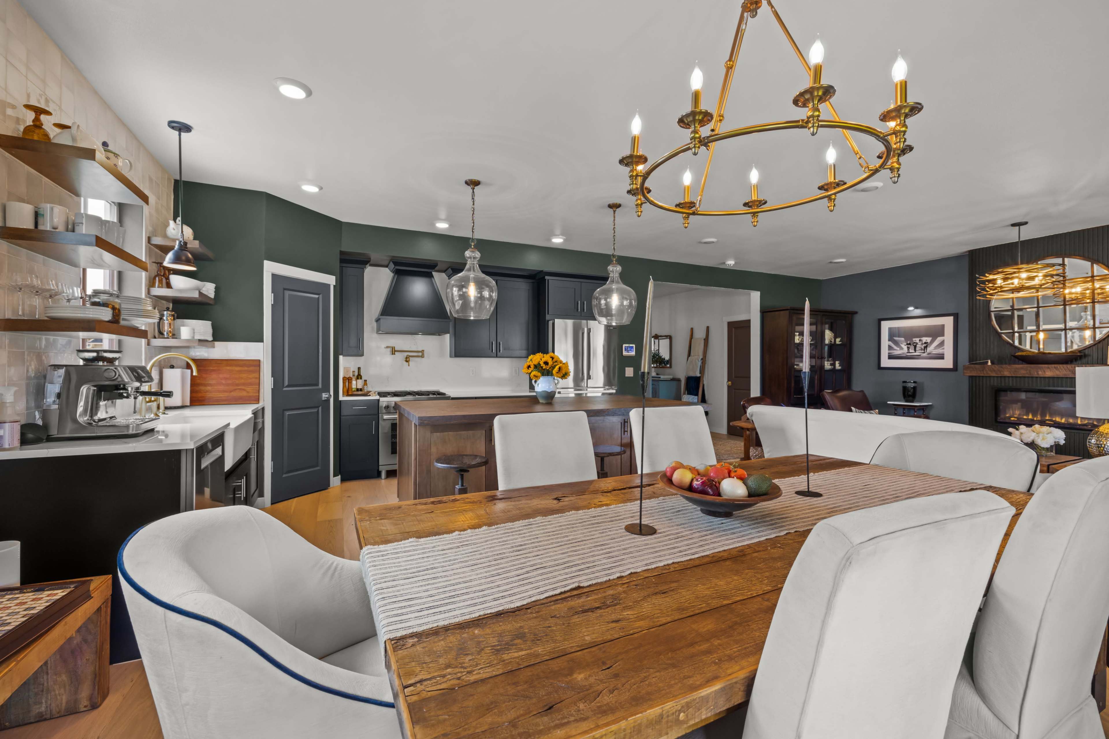 A dining table set with a bowl of fruit is positioned in a modern kitchen and dining area featuring pendant lighting and a cozy seating arrangement.