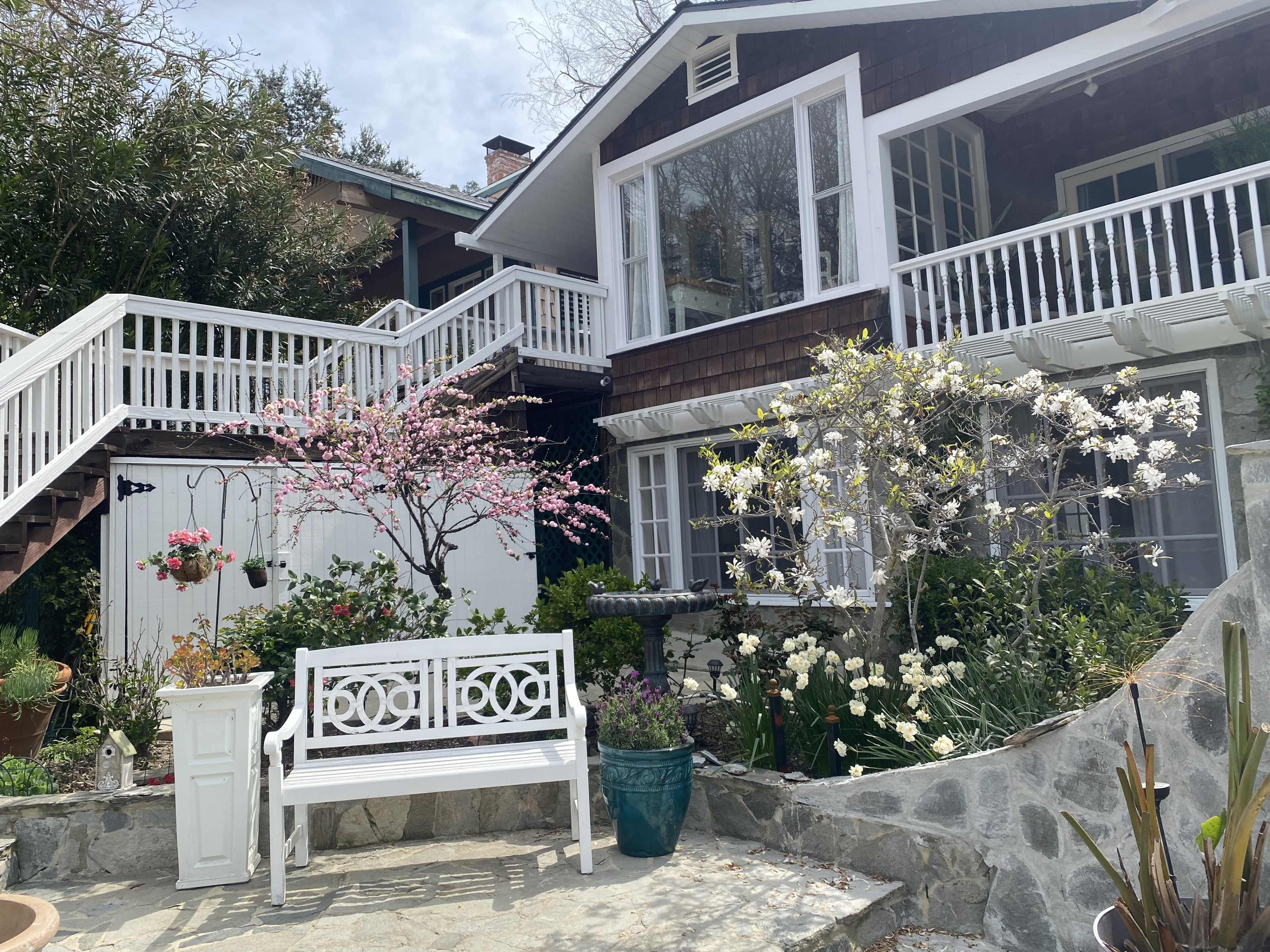 A two-story house with a wooden exterior features a garden with blooming trees, flowers, and a white bench.