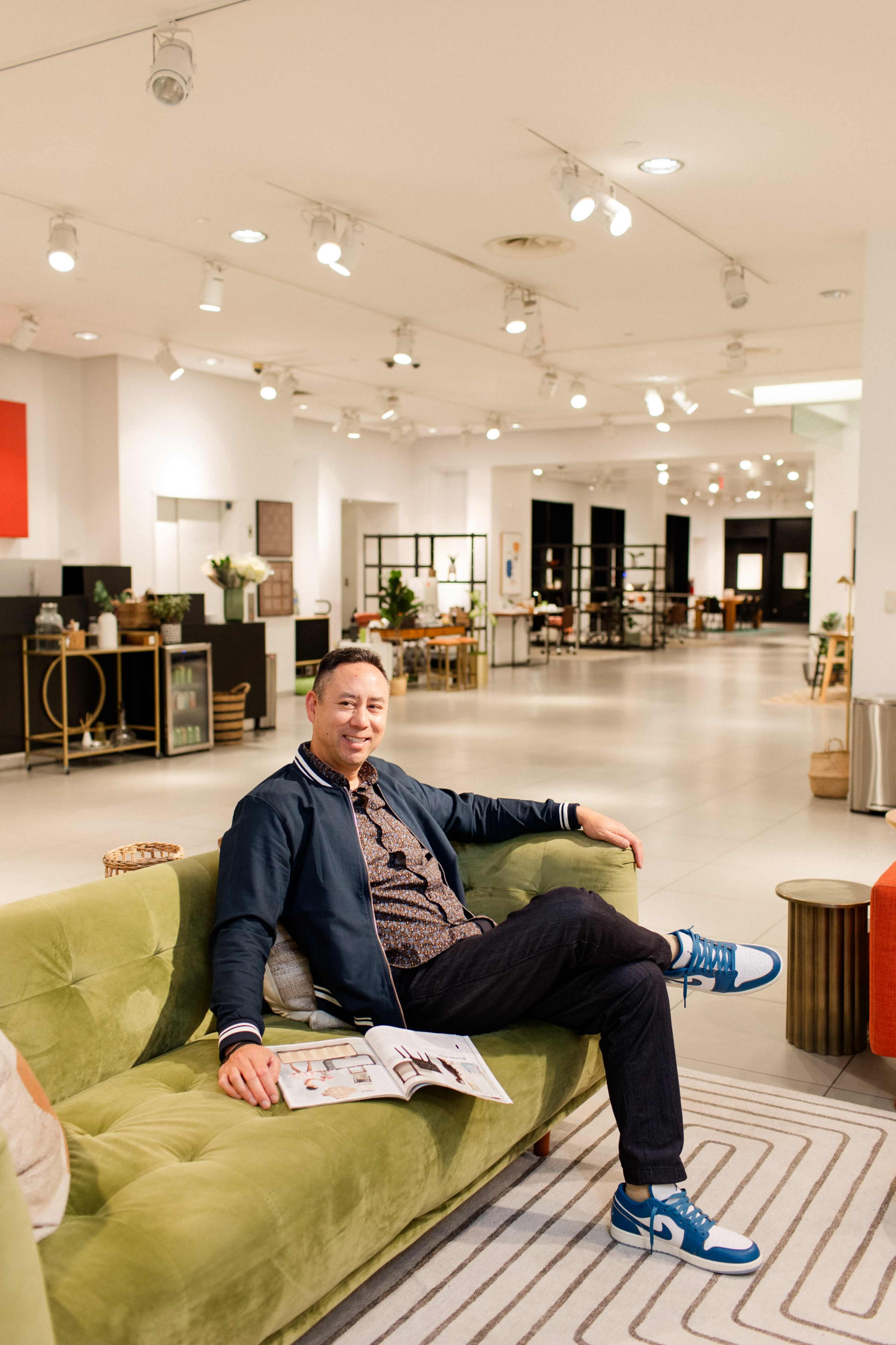 A man sits on a green couch in a well-lit modern furniture showroom, holding a magazine.