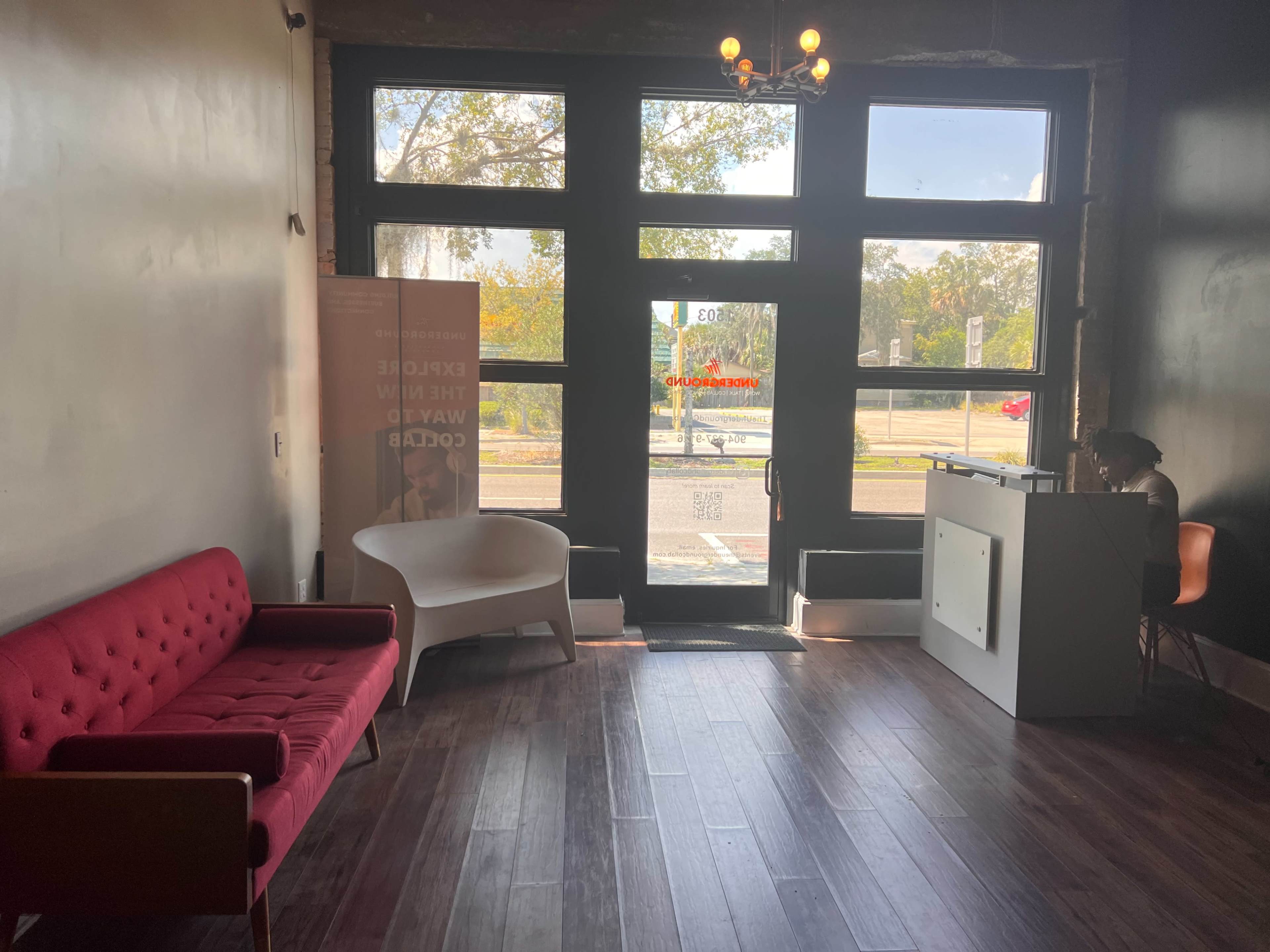 The interior of a modern waiting area features a red couch, a white chair, and a reception desk near large windows that overlook the street.