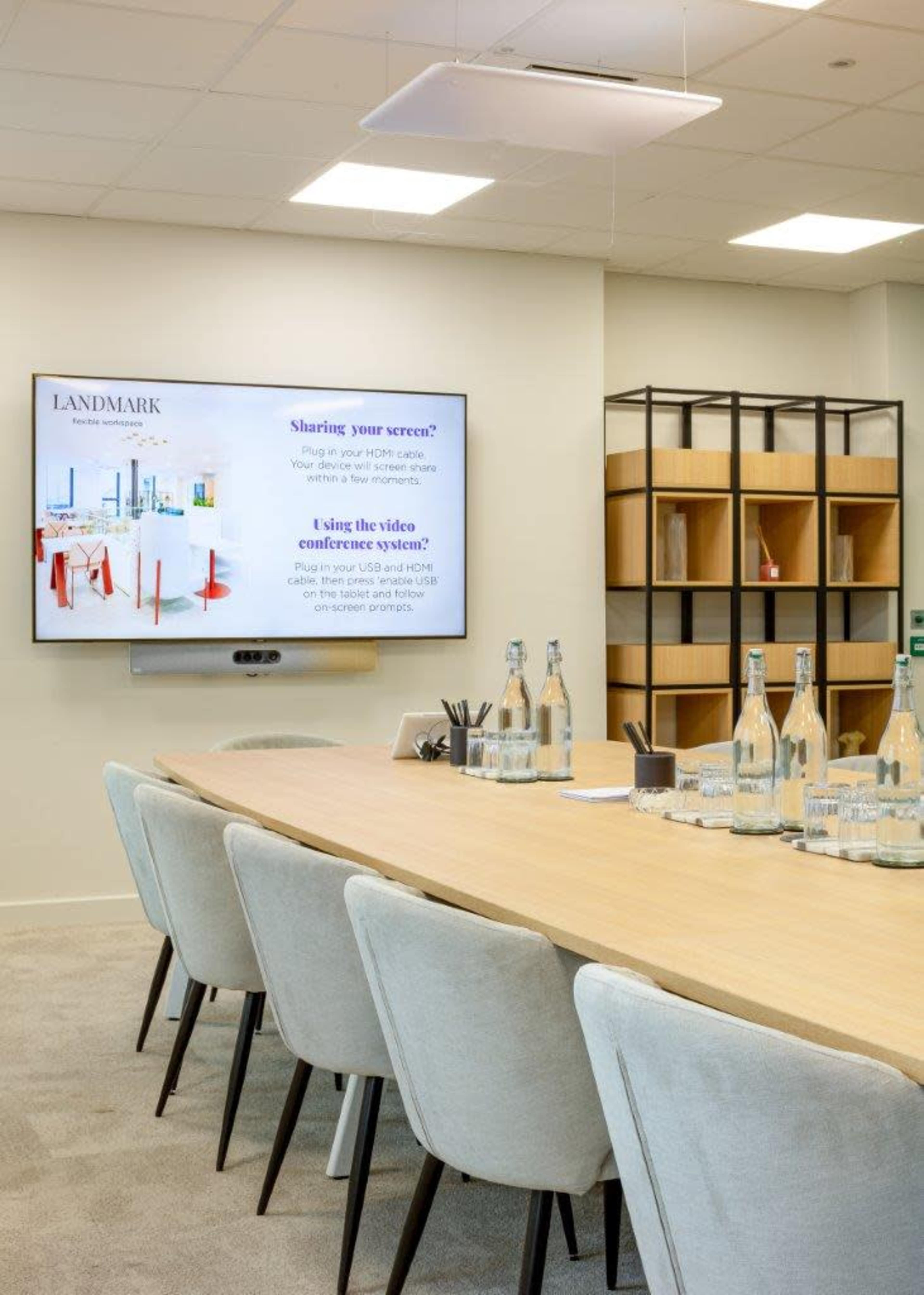 The image shows a modern conference room with a long wooden table, ergonomic chairs, a television display on the wall, and shelves filled with organizational materials.