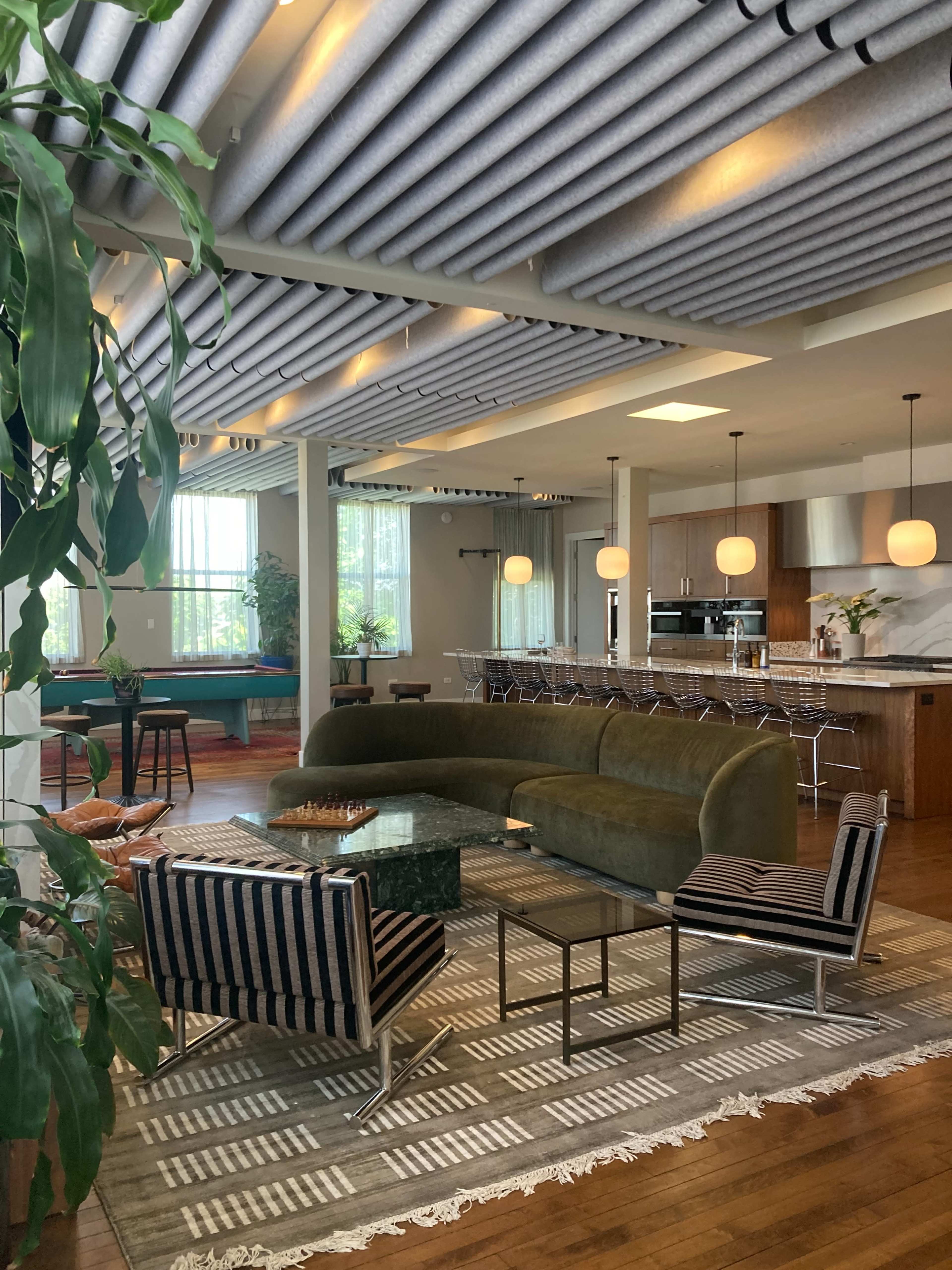 The image shows a modern living space featuring a green curved sofa, striped chairs, a glass coffee table, and an open kitchen with stylish lighting fixtures.