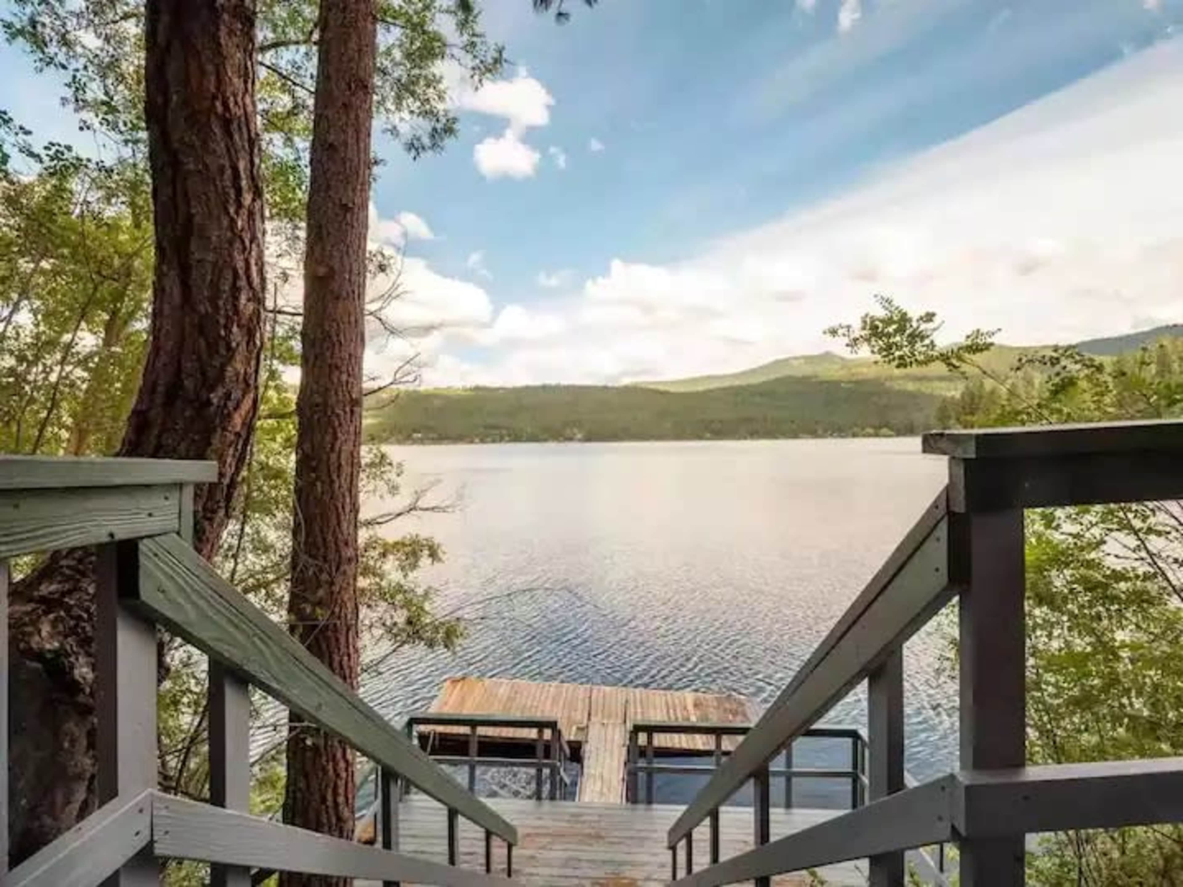 A wooden dock extends over a calm lake surrounded by trees and mountains under a partly cloudy sky.