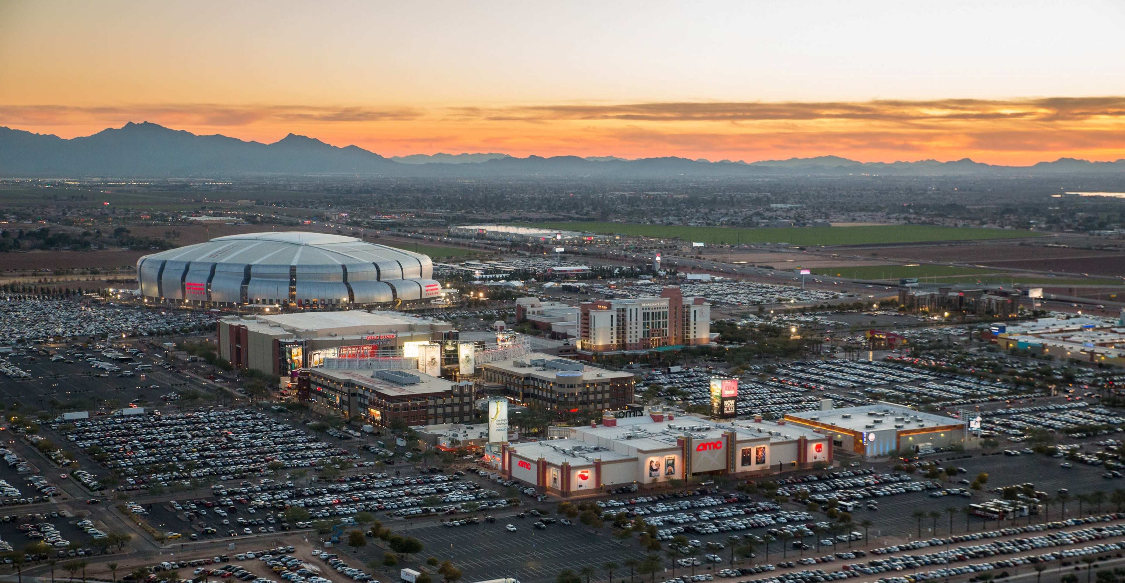 A large stadium next to a commercial complex with multiple buildings and a vast parking lot during sunset.