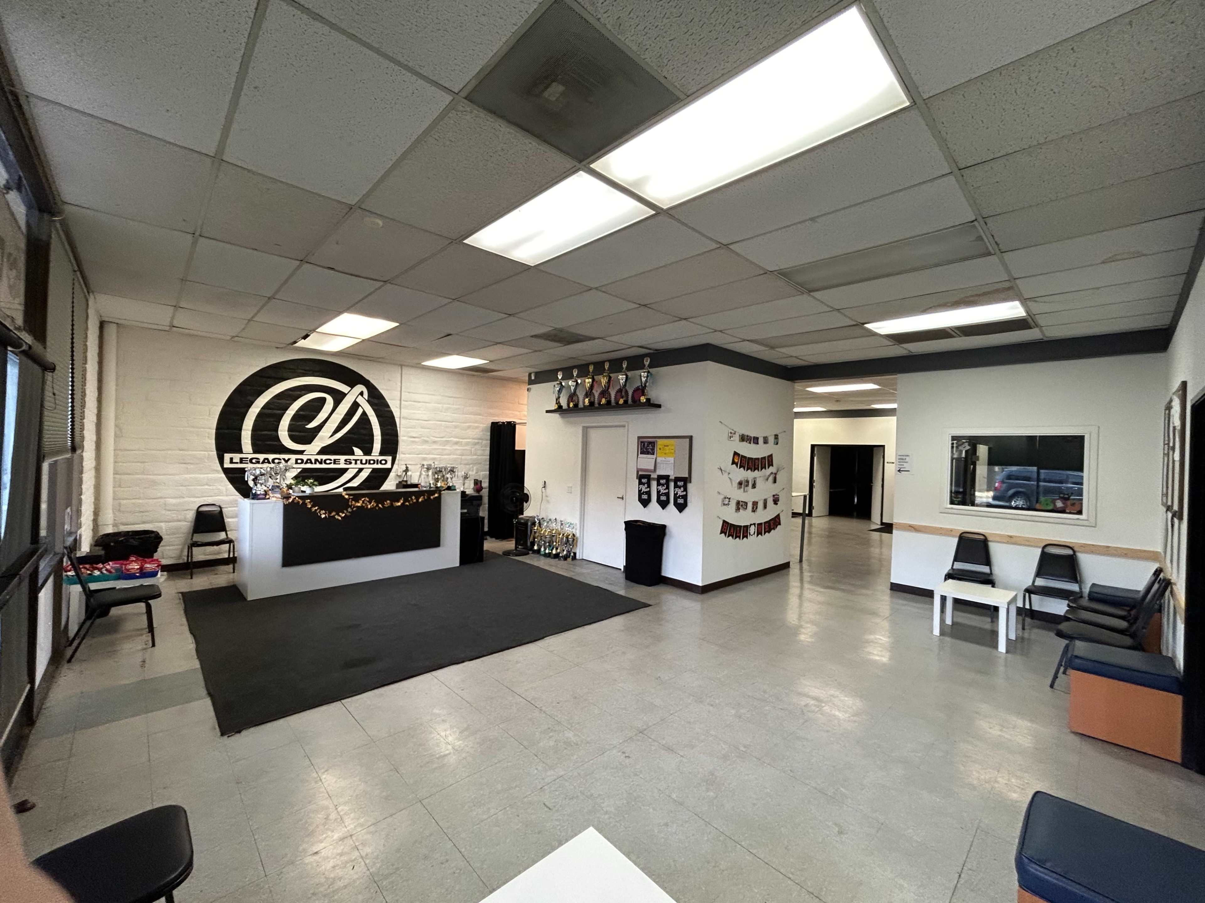 The image shows the interior of a dance studio with a reception area, a black dance floor, and seating along the walls.