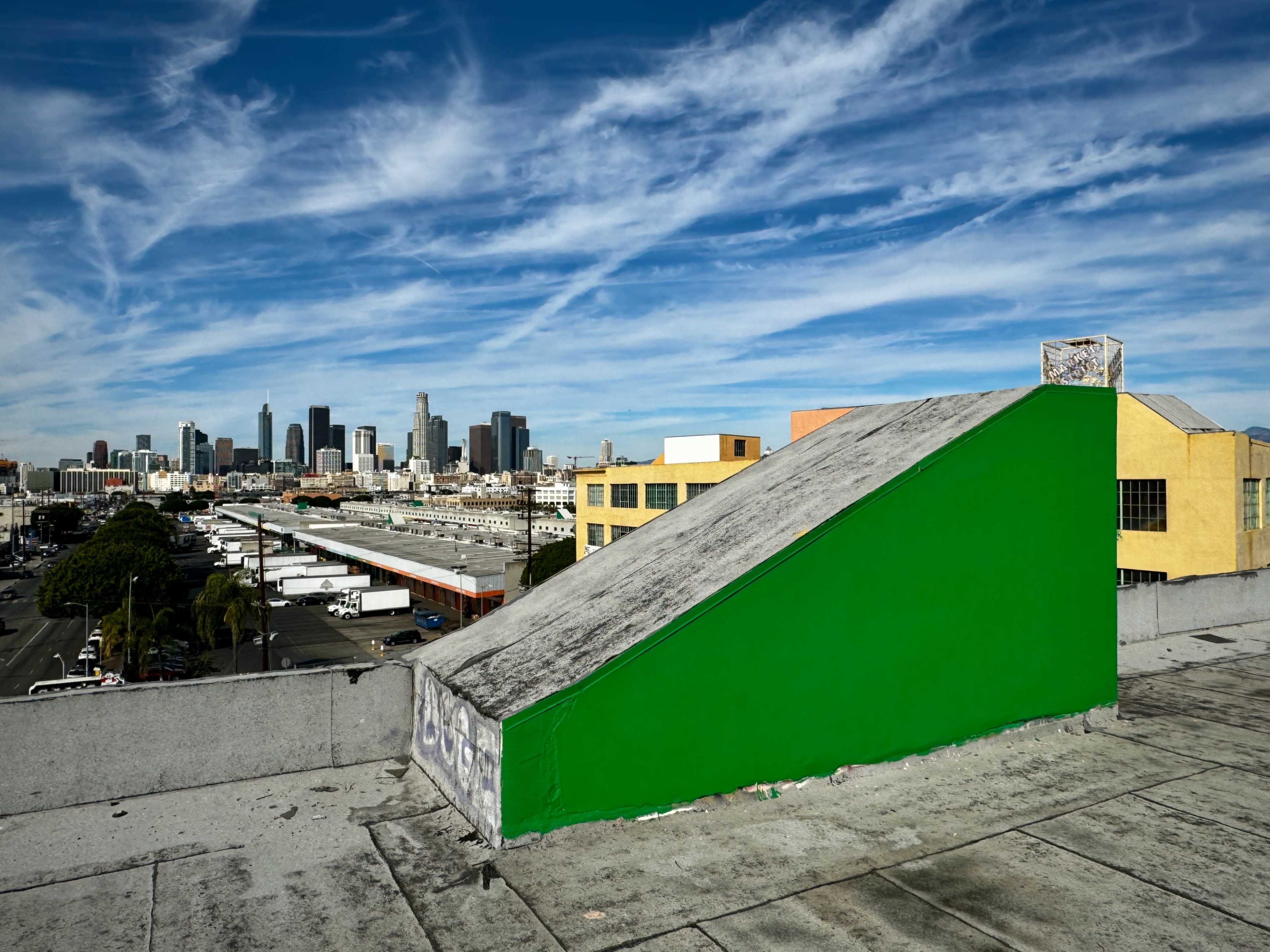 A bright green structure on a rooftop stands against a backdrop of downtown skyscrapers and a blue sky with clouds.