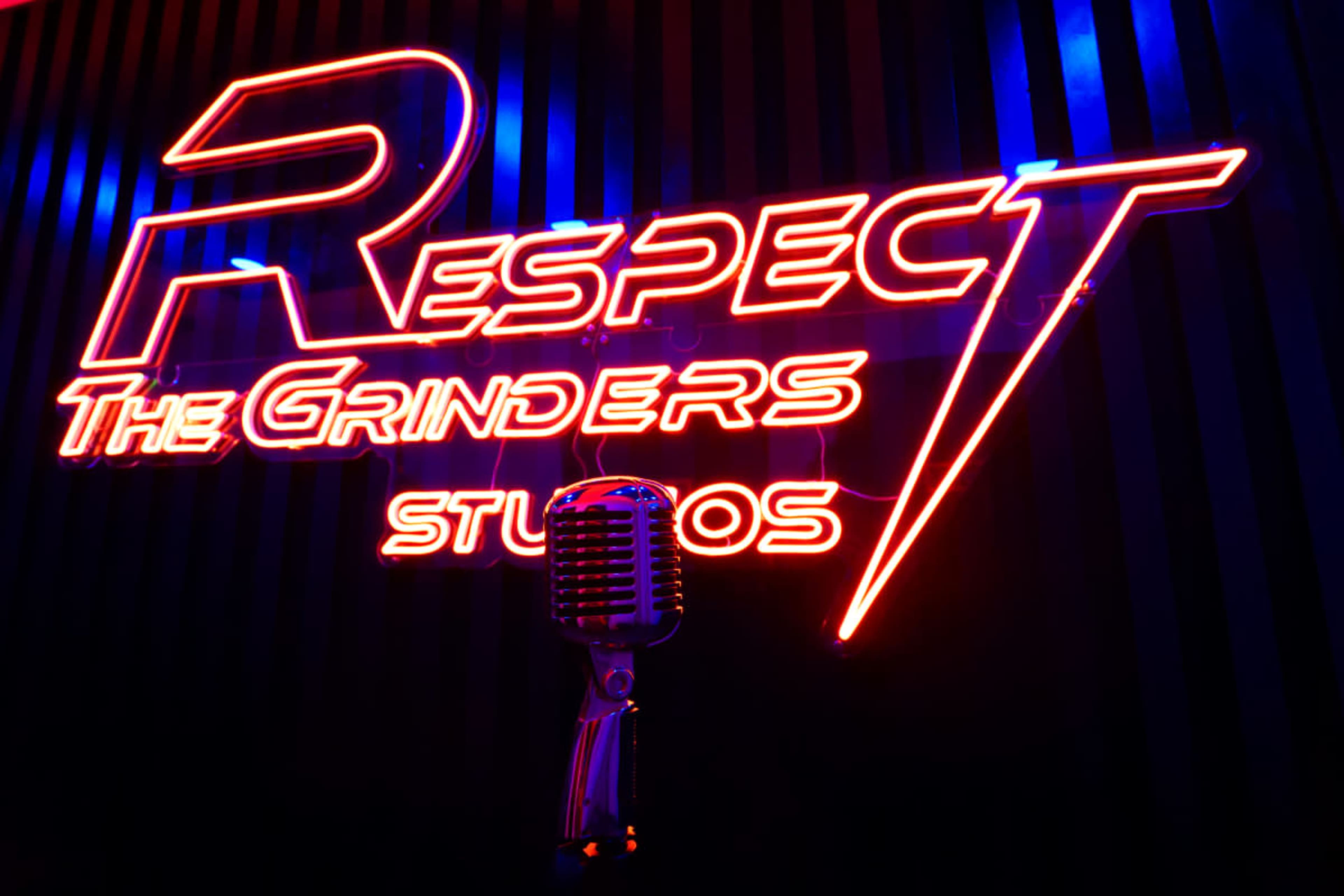 The image features a vibrant neon sign that reads "Respect The Grinders" above a vintage microphone mounted on a wall.