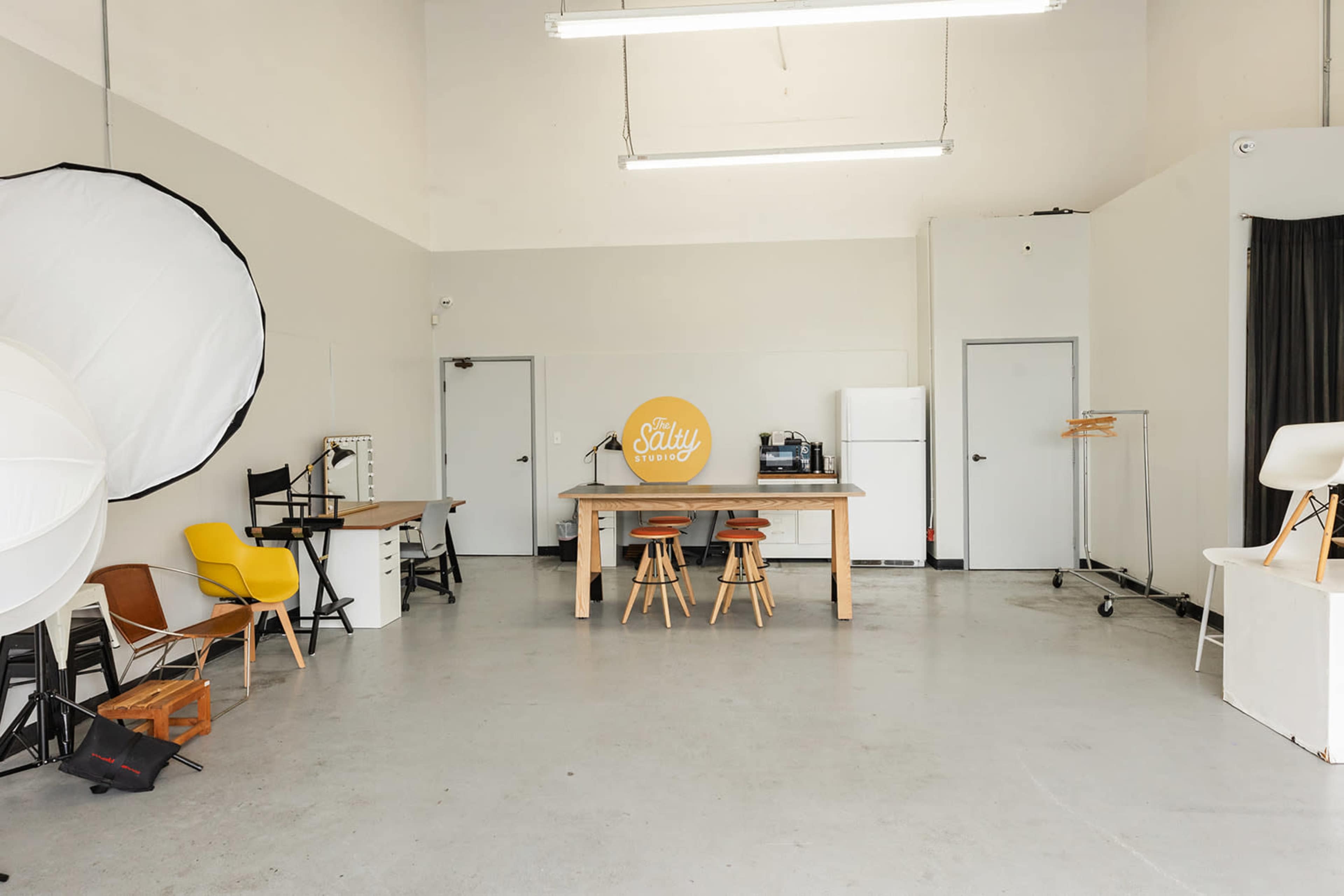 A spacious studio features a large table with three stools, various chairs, a refrigerator, and softbox lighting equipment.