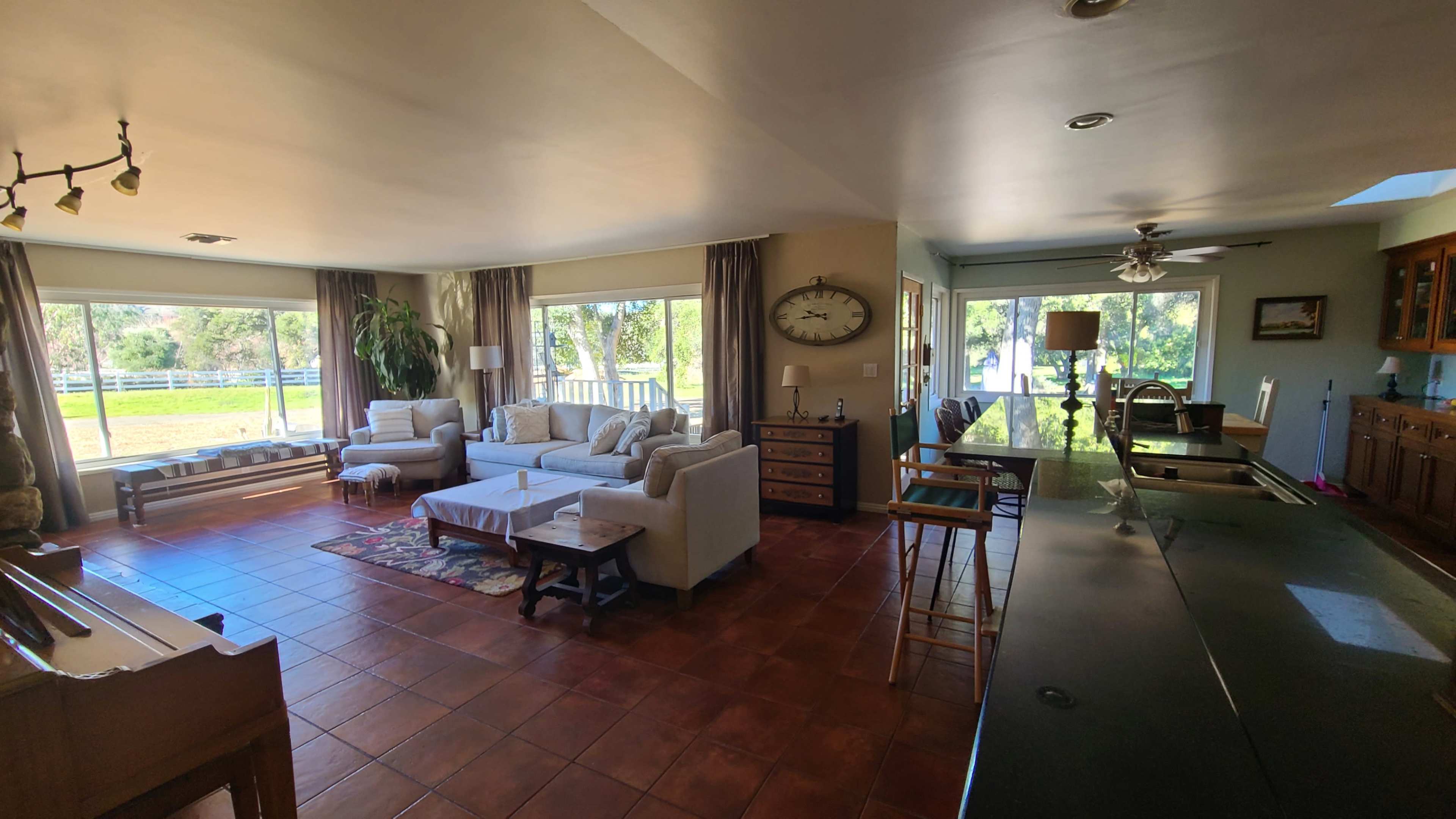 The image shows a spacious living room with several seating areas, large windows providing natural light, and a view of an outdoor space.