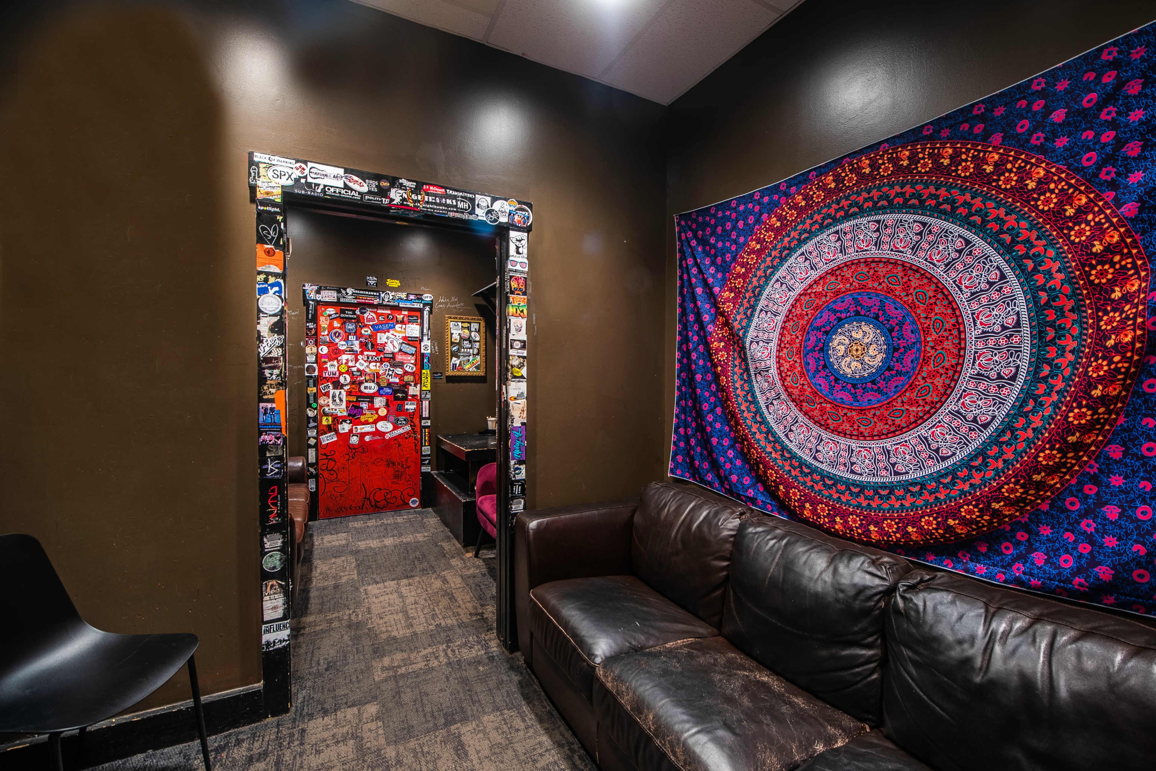 The image shows a cozy, dimly lit room featuring a dark brown wall, a patterned tapestry, a black couch, and a doorway decorated with colorful stickers.