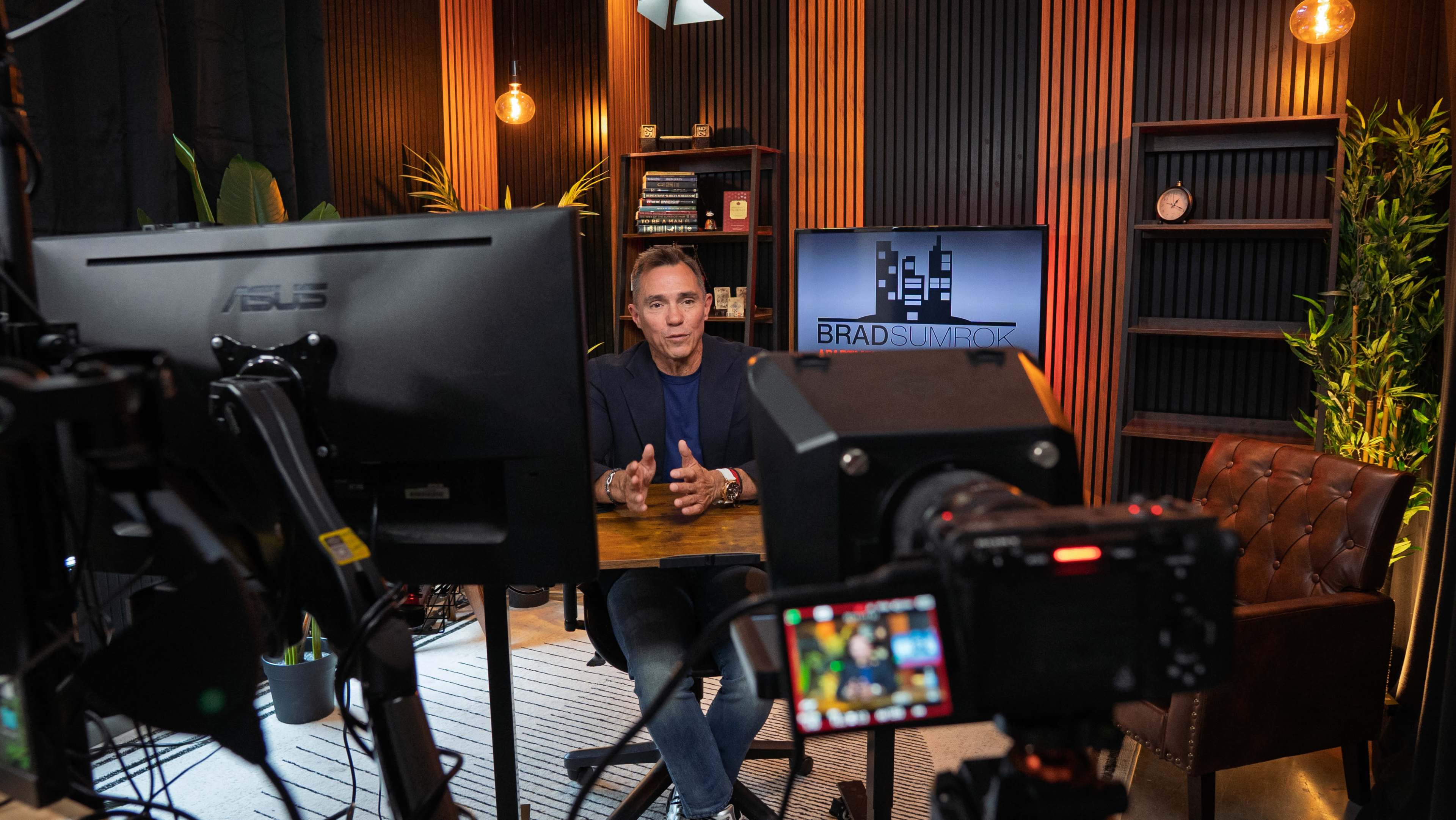 A man sits at a desk in a well-decorated office space, speaking into a camera while a computer monitor displays a logo in the background.