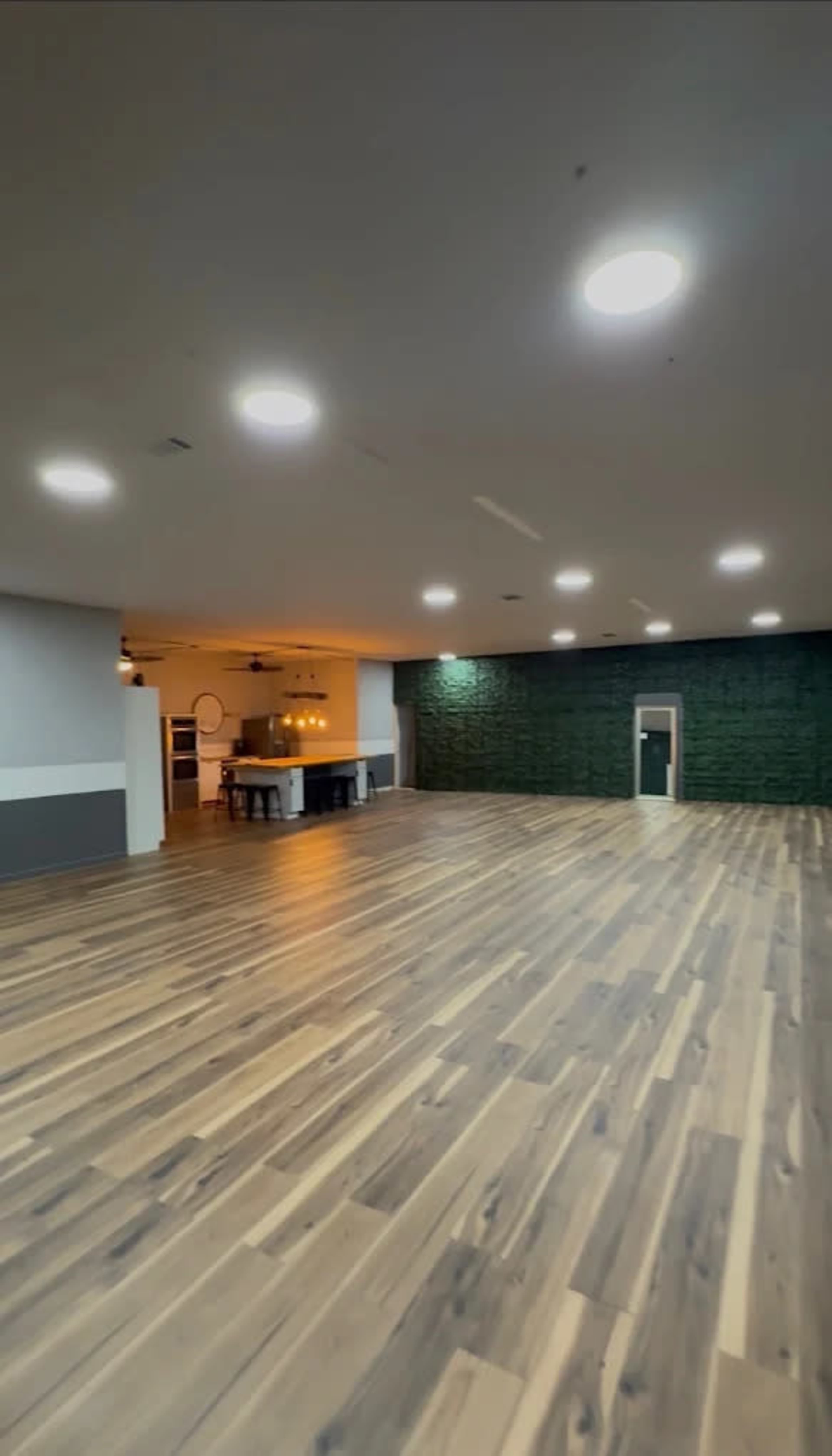 The image shows a spacious empty room with wooden flooring, featuring a wall with green decor and a doorway on one side leading to a kitchen area.