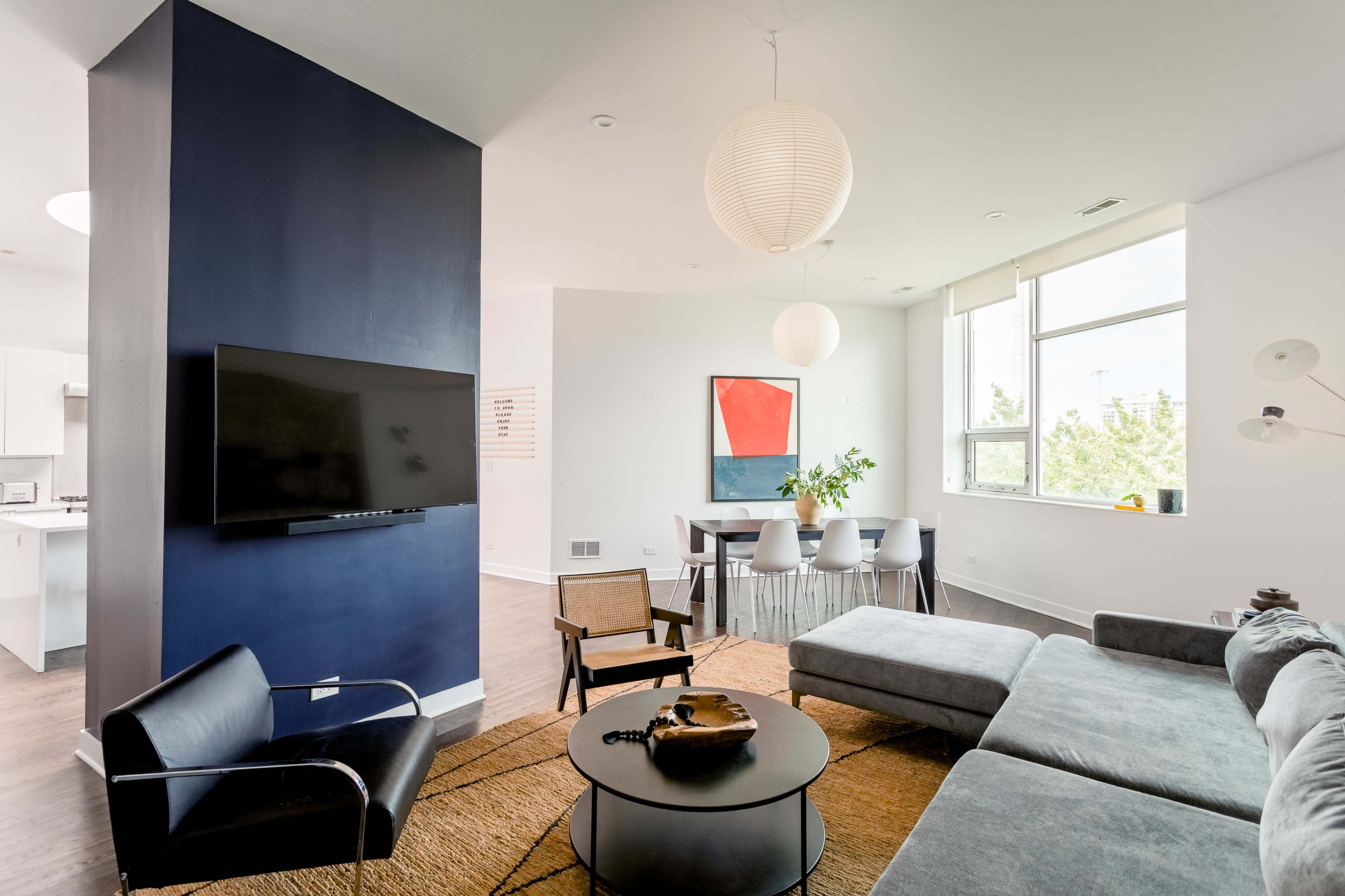A modern living space featuring a sectional sofa, a round coffee table, a wall-mounted television, and a dining area with a table and chairs, complemented by a large window and minimalist decor.