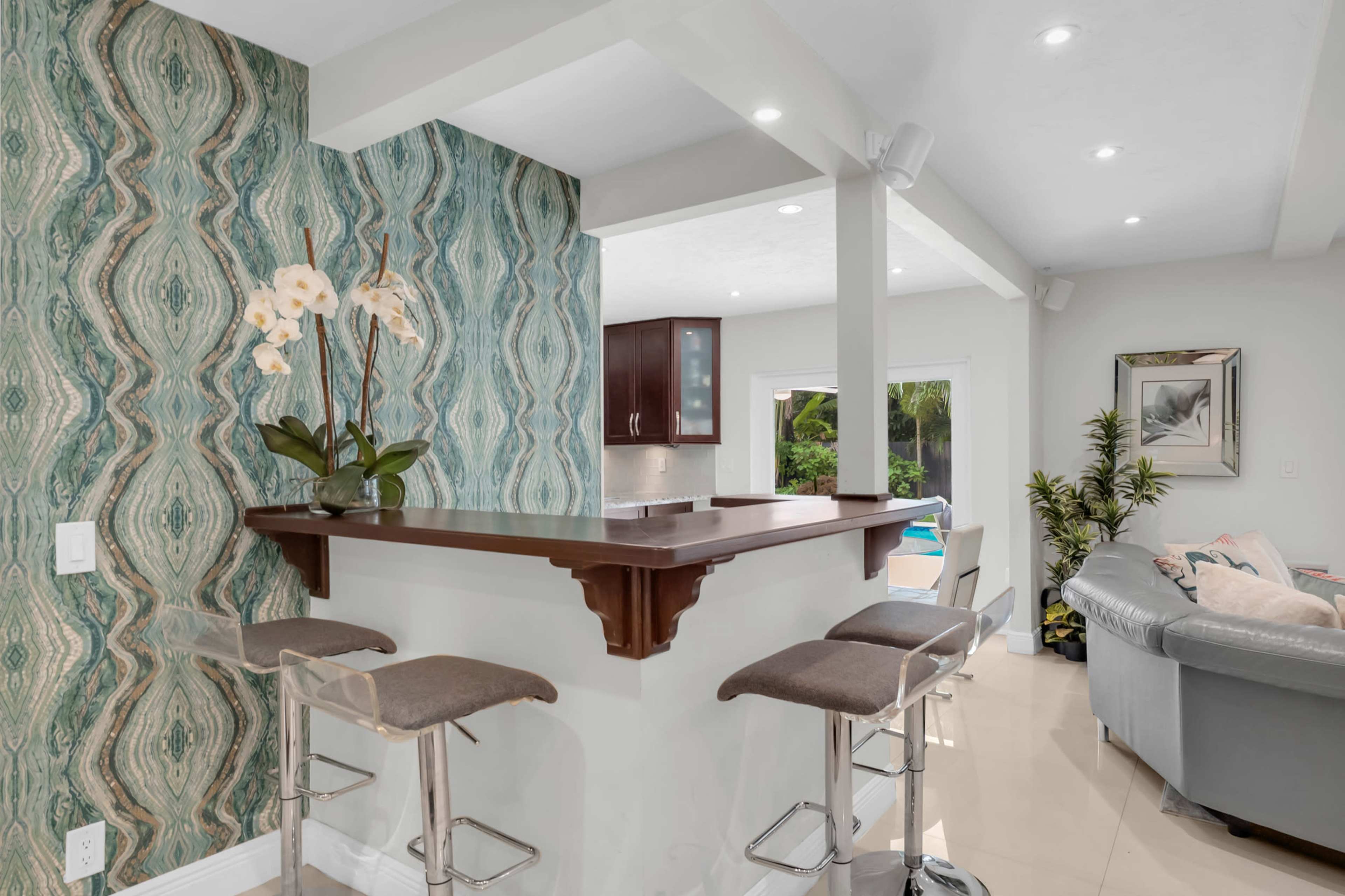 The image shows a modern kitchen area with a bar counter featuring four metal stools, surrounded by decorative wallpaper and cabinetry.