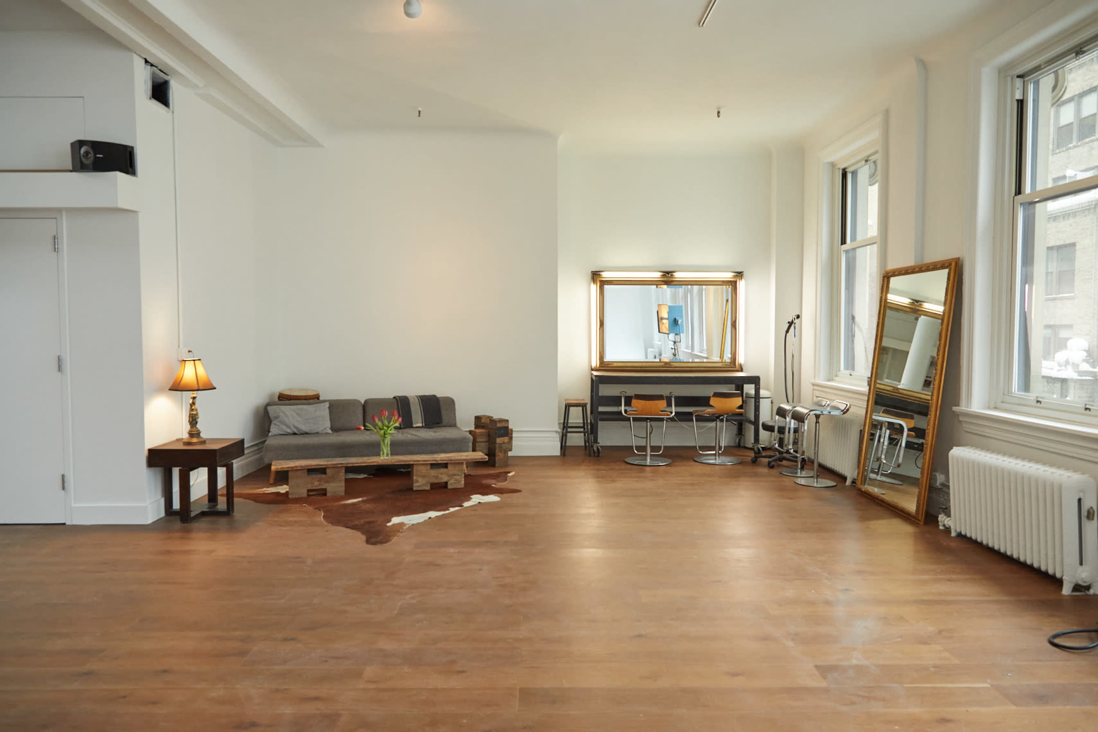 A spacious, well-lit room features a gray couch, a wooden coffee table, a vanity mirror with lights, and two bar stools along with a radiator against the wall.