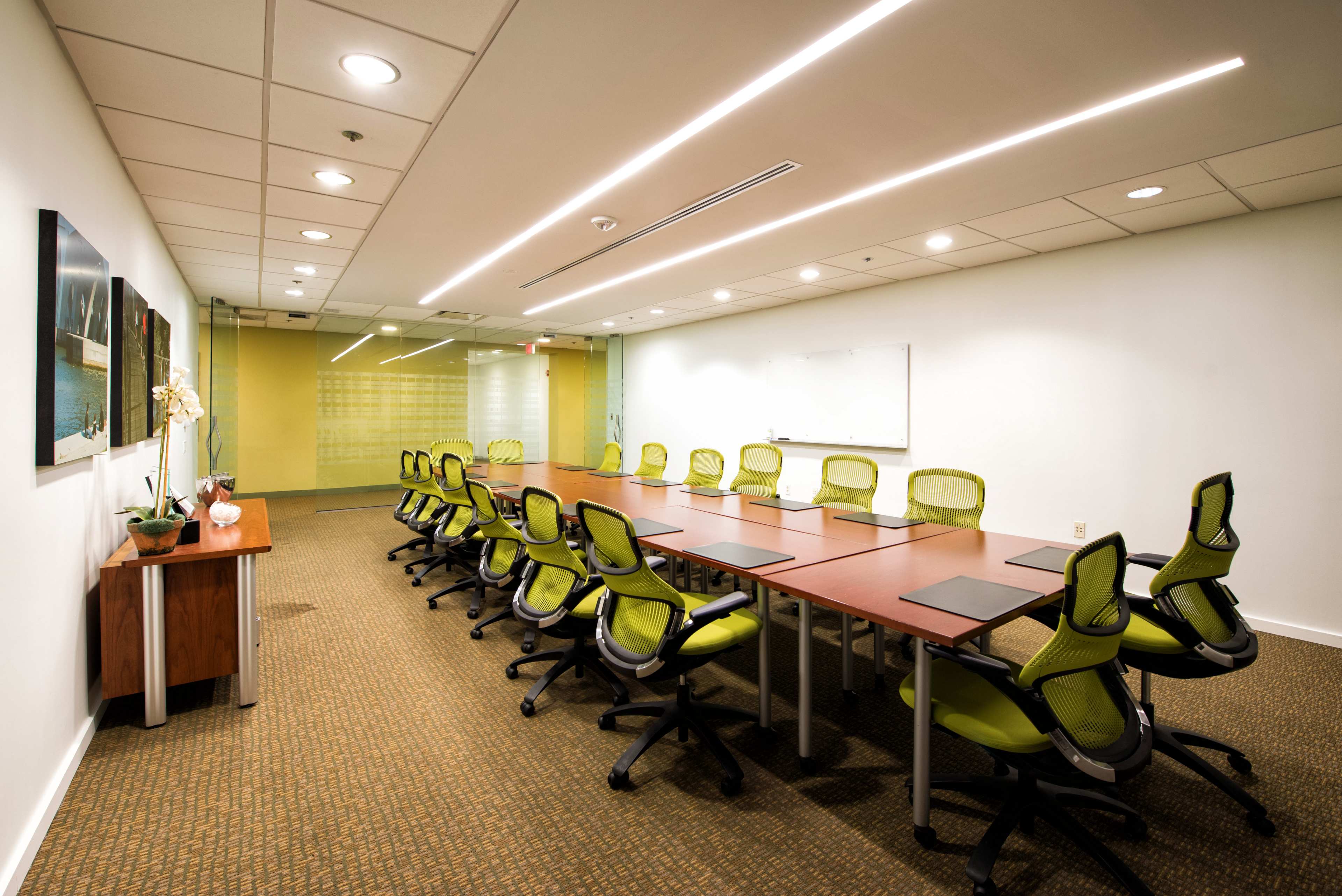 A conference room features a long rectangular table with green chairs arranged around it, and a whiteboard is mounted on the wall opposite a glass panel.
