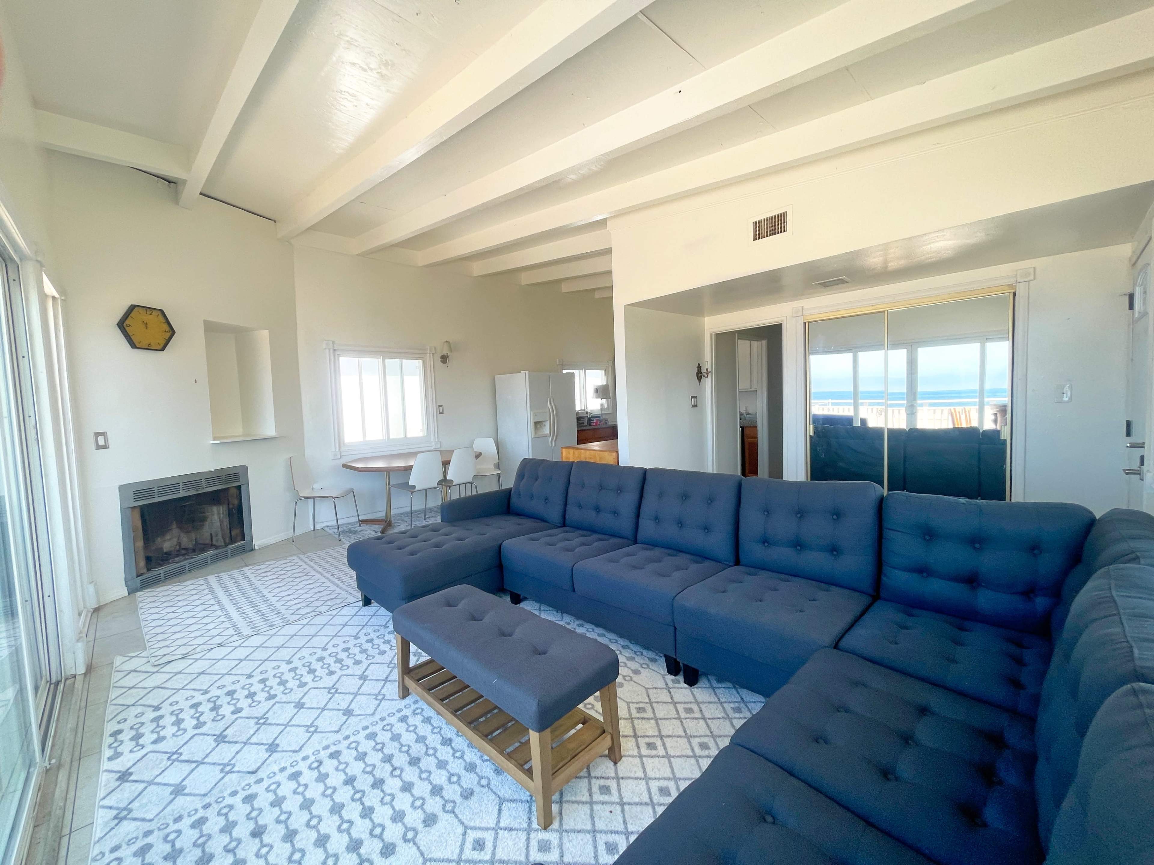 A spacious living room featuring a large blue sectional sofa, a fireplace, and a dining area with a table and chairs, illuminated by natural light from large windows.