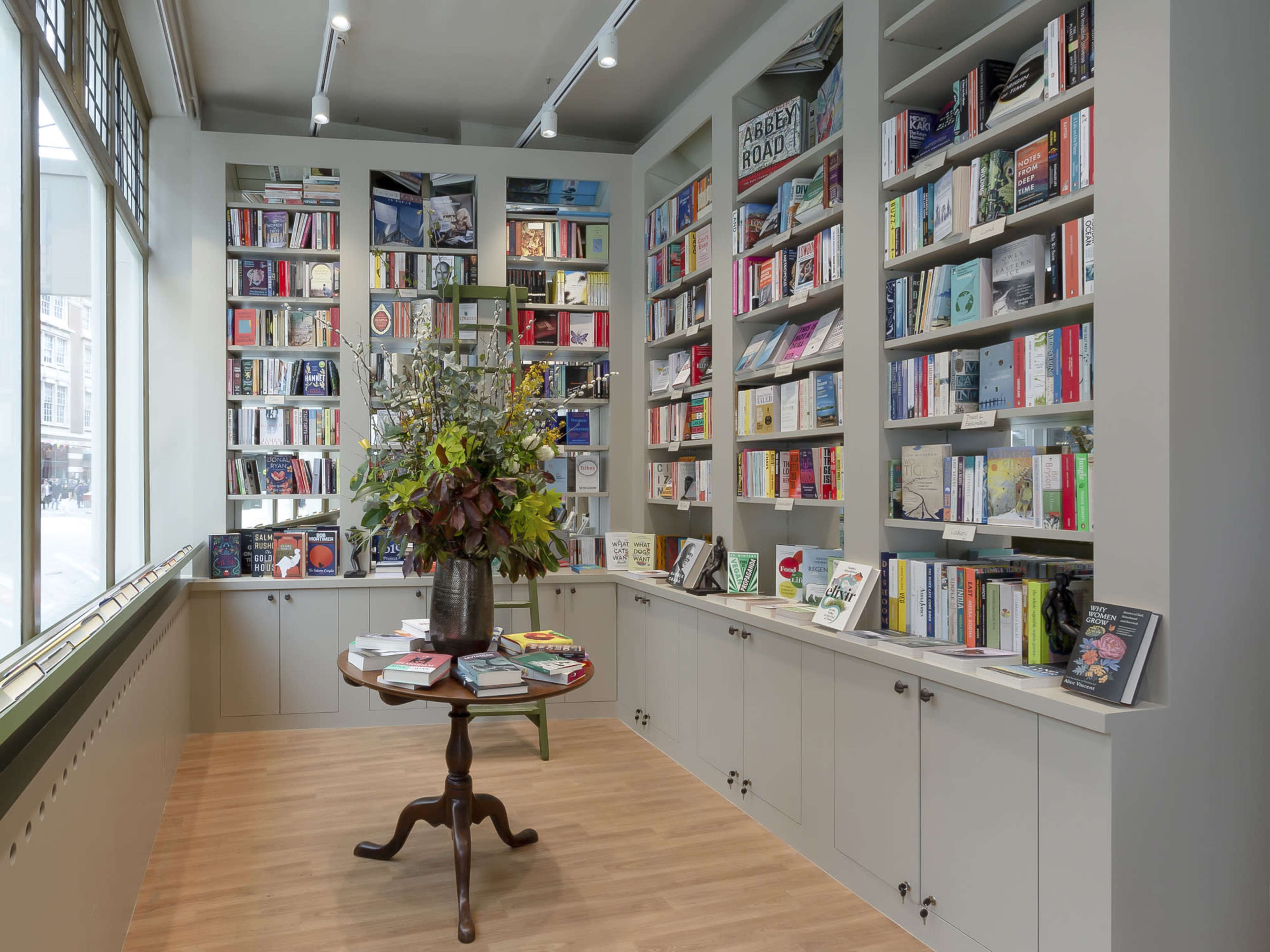 A cozy bookstore interior with shelves filled with books, a central round table adorned with a floral arrangement, and large windows allowing natural light to enter.