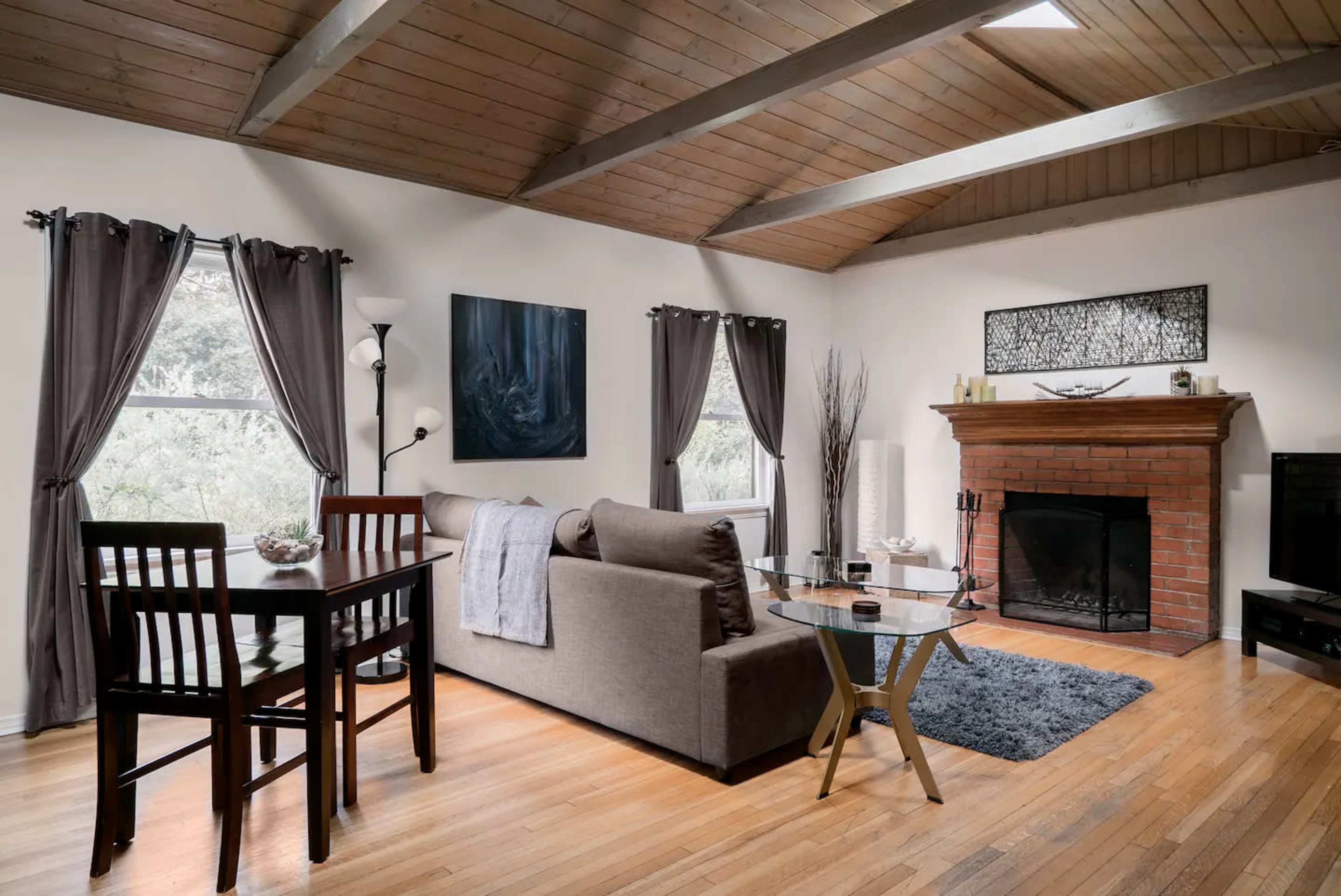 The image shows a living room with a gray sofa, a glass coffee table, a dining area with high stools, and a fireplace surrounded by a wooden beam ceiling.