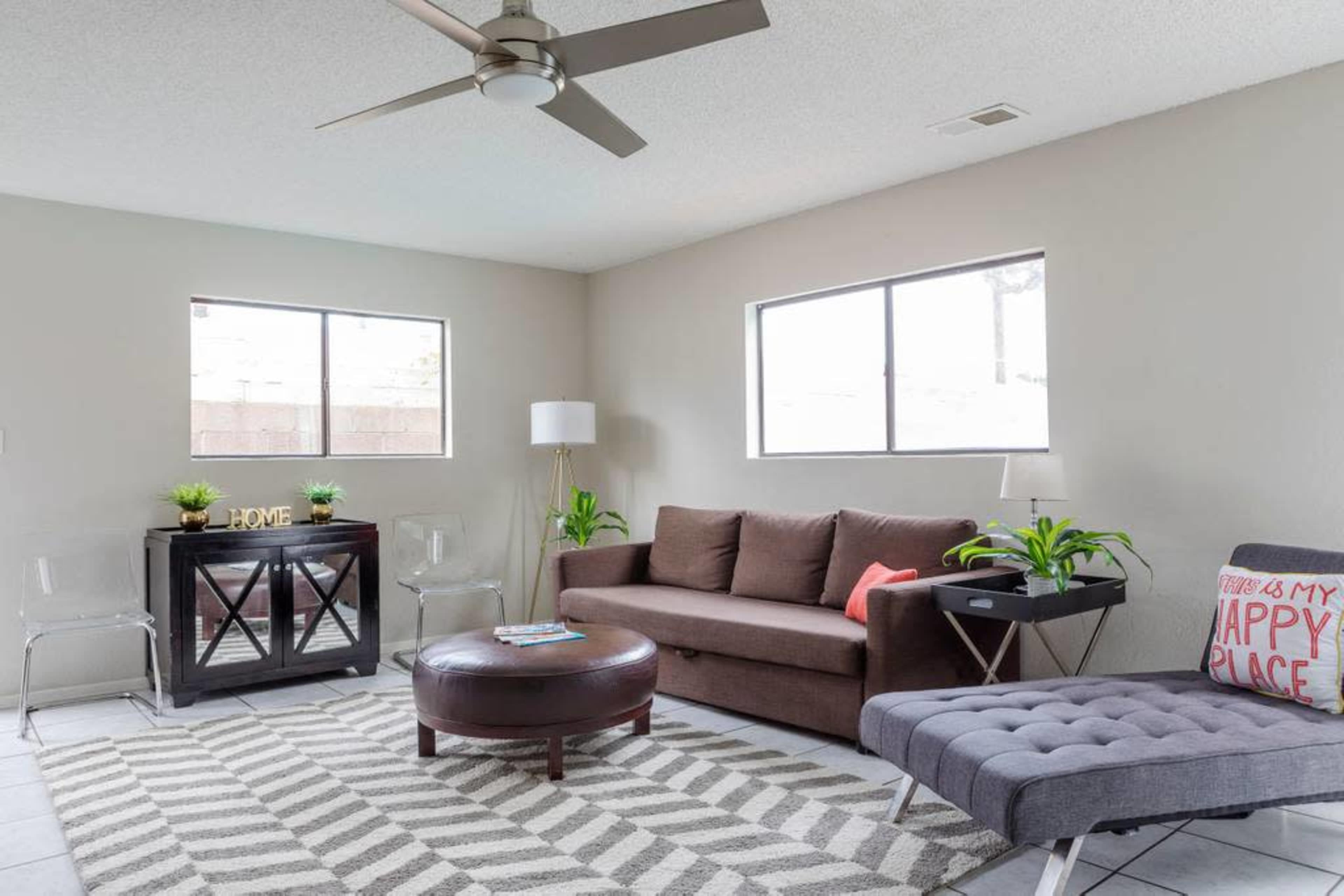 A living room features a brown sofa, a gray chaise lounge, a glass side table, and plants, with two windows allowing natural light.