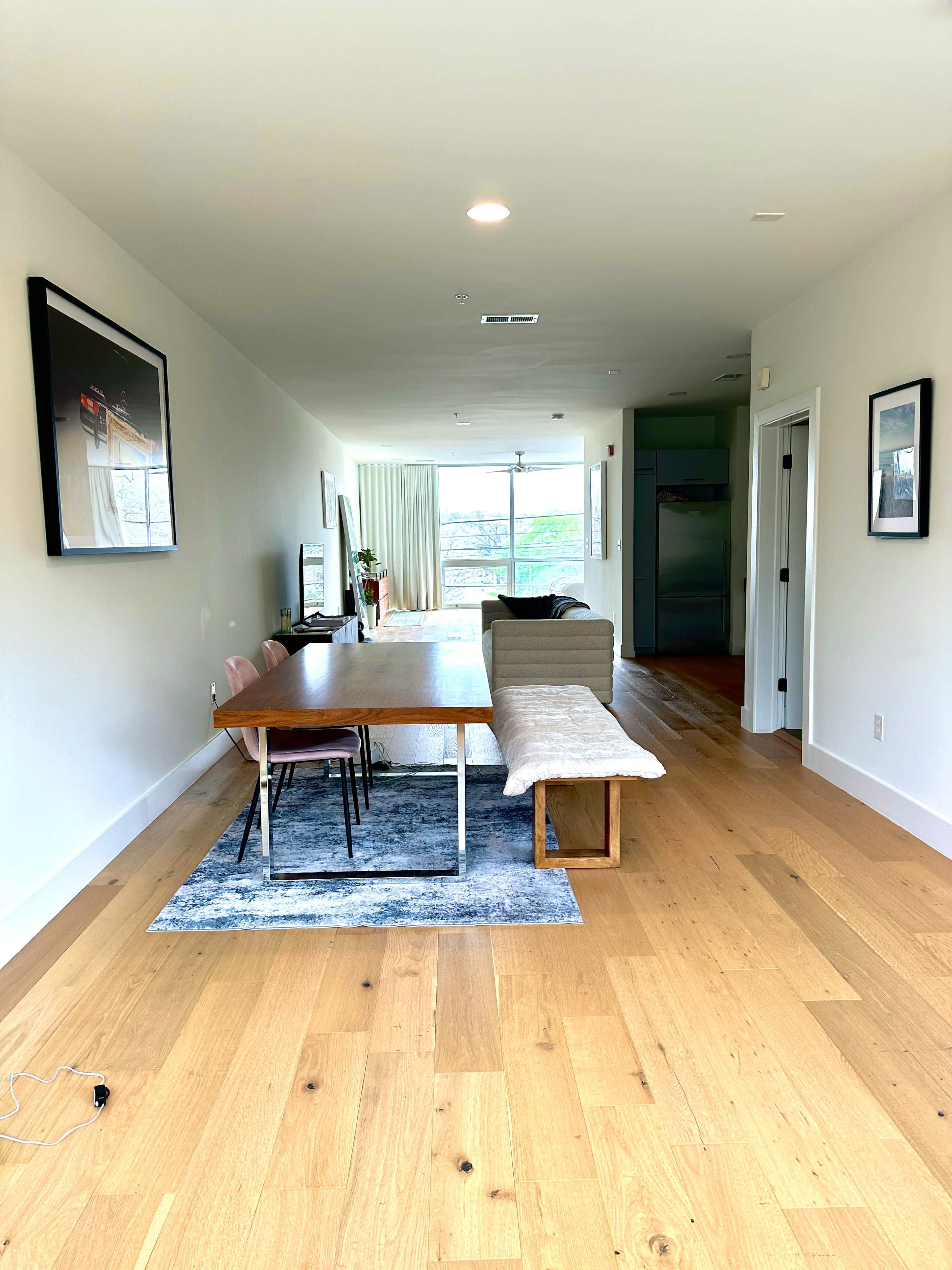 A spacious living area features a wooden dining table with chairs, a couch, and large windows allowing natural light to enter.