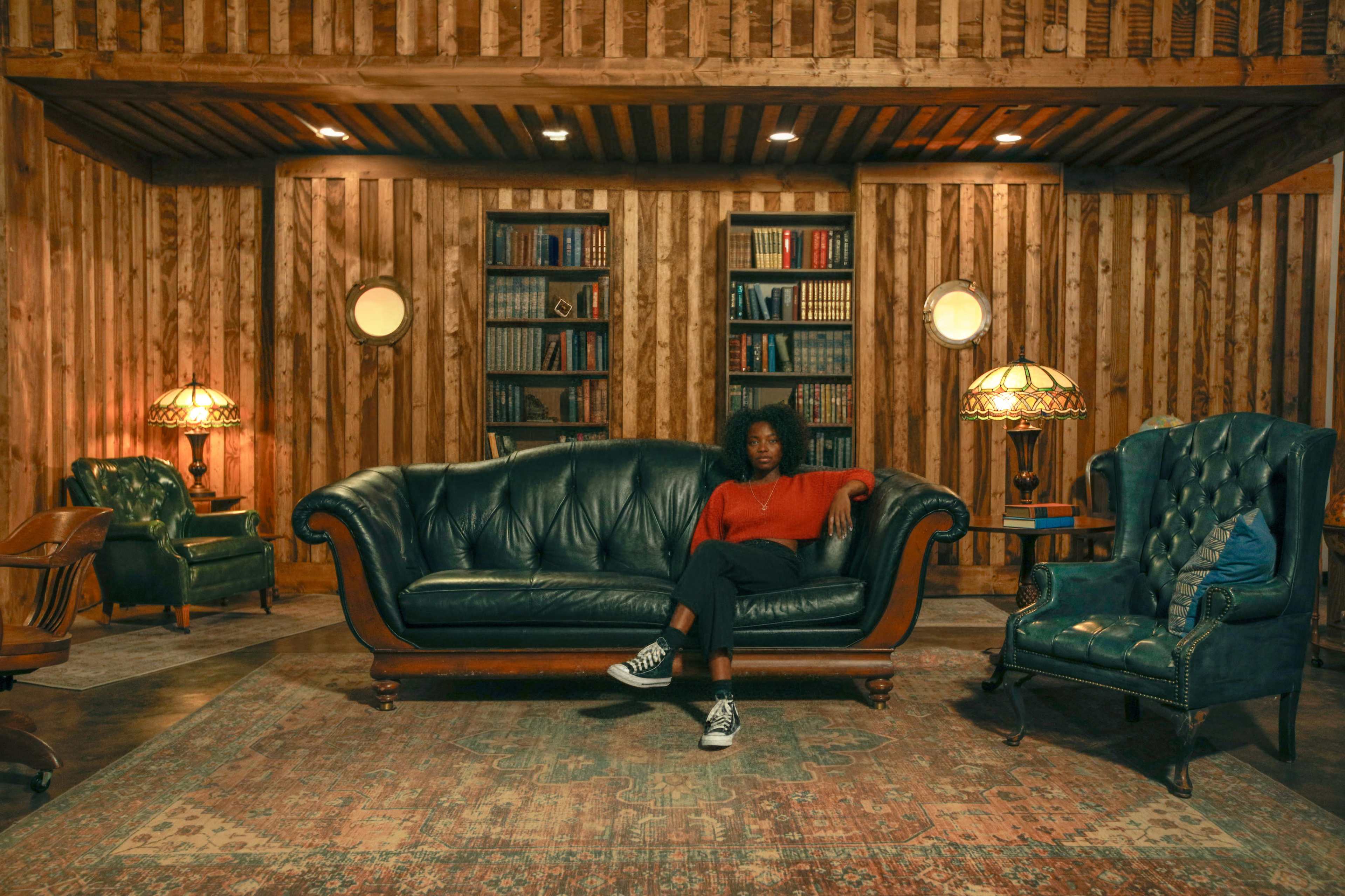 A person sits on a dark green leather couch in a room with wooden walls and bookshelves, illuminated by two lamps.