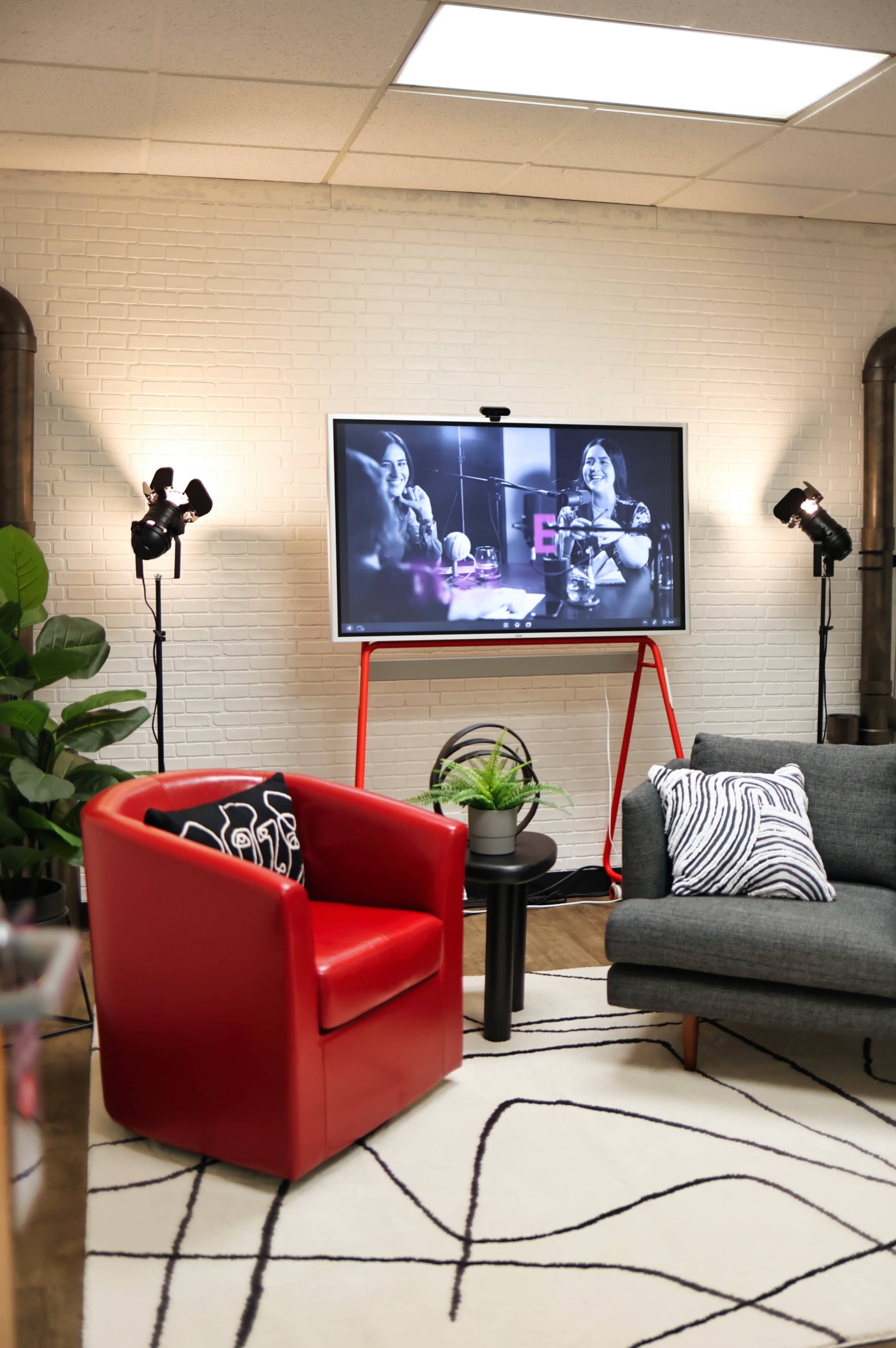 A cozy lounge area features a red armchair, a gray sofa, and a television displaying a black-and-white video, surrounded by plants and studio lights.