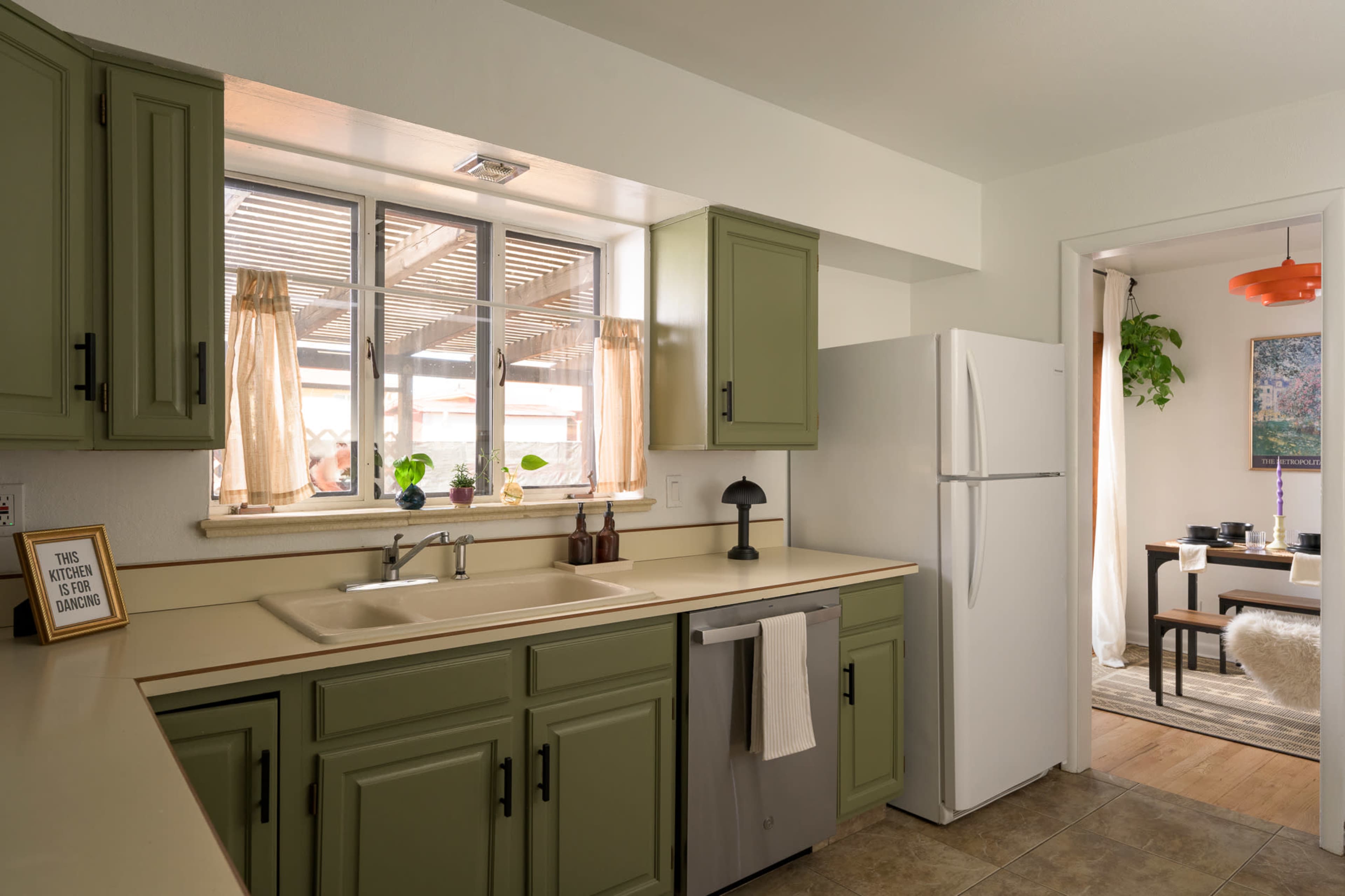 A kitchen features olive green cabinets, a window with sheer curtains, and a white refrigerator, with a view into a dining area beyond.