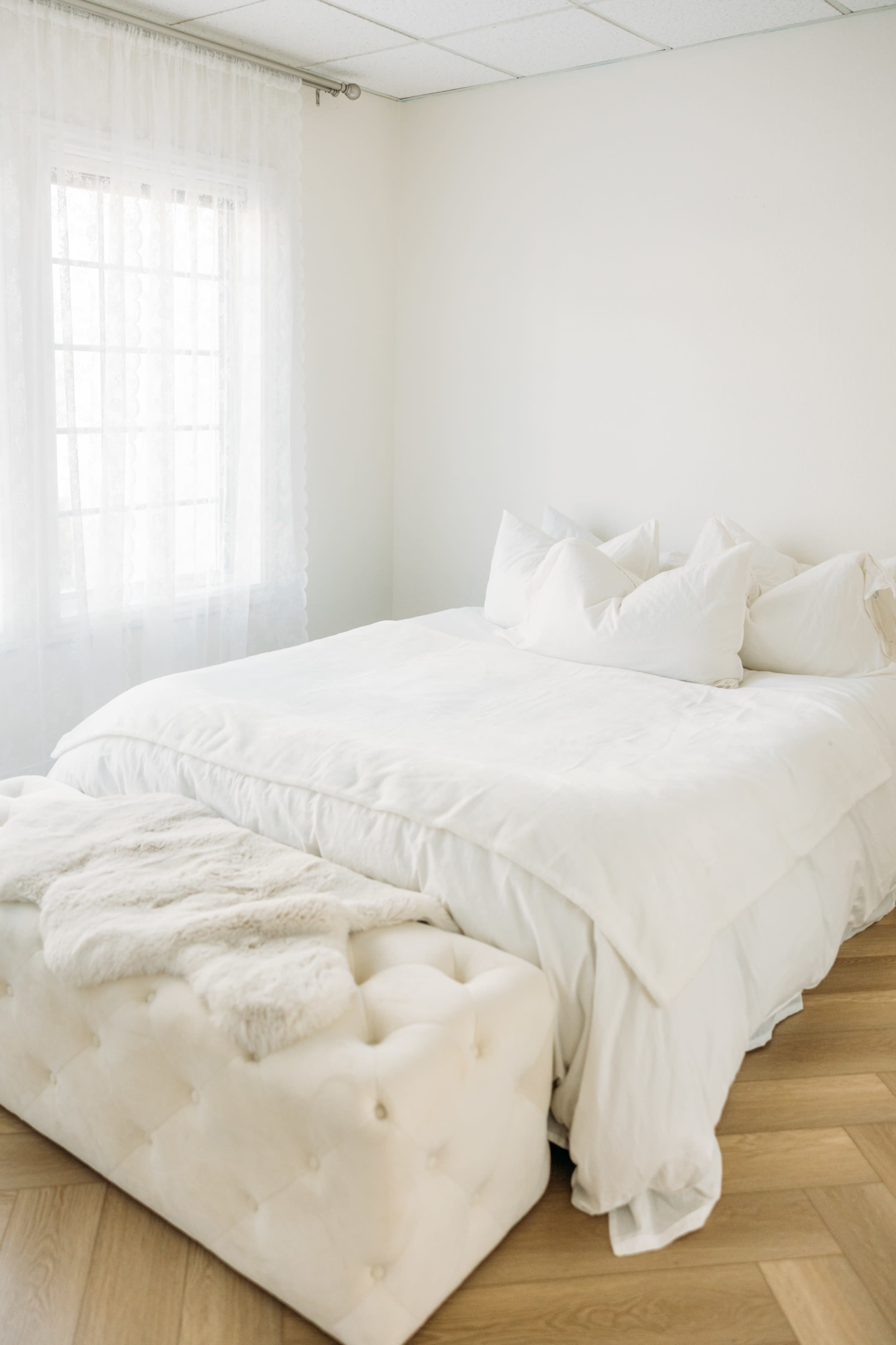 A neatly made bed with white bedding is positioned beside a tufted ottoman in a bright, minimalist room.