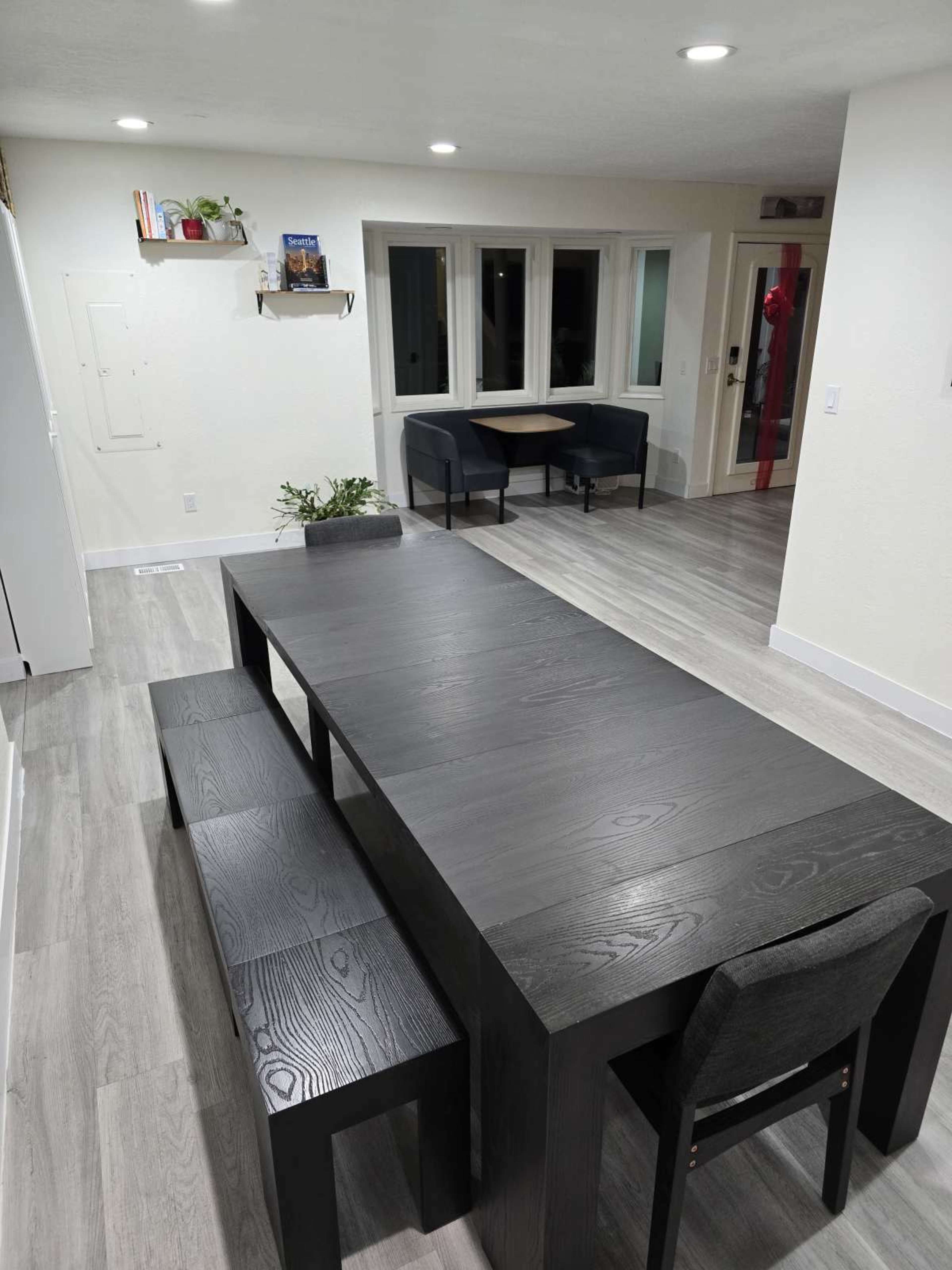 The image shows a modern dining area featuring a large black table with a bench and a chair, set in a well-lit room with a minimalist design.