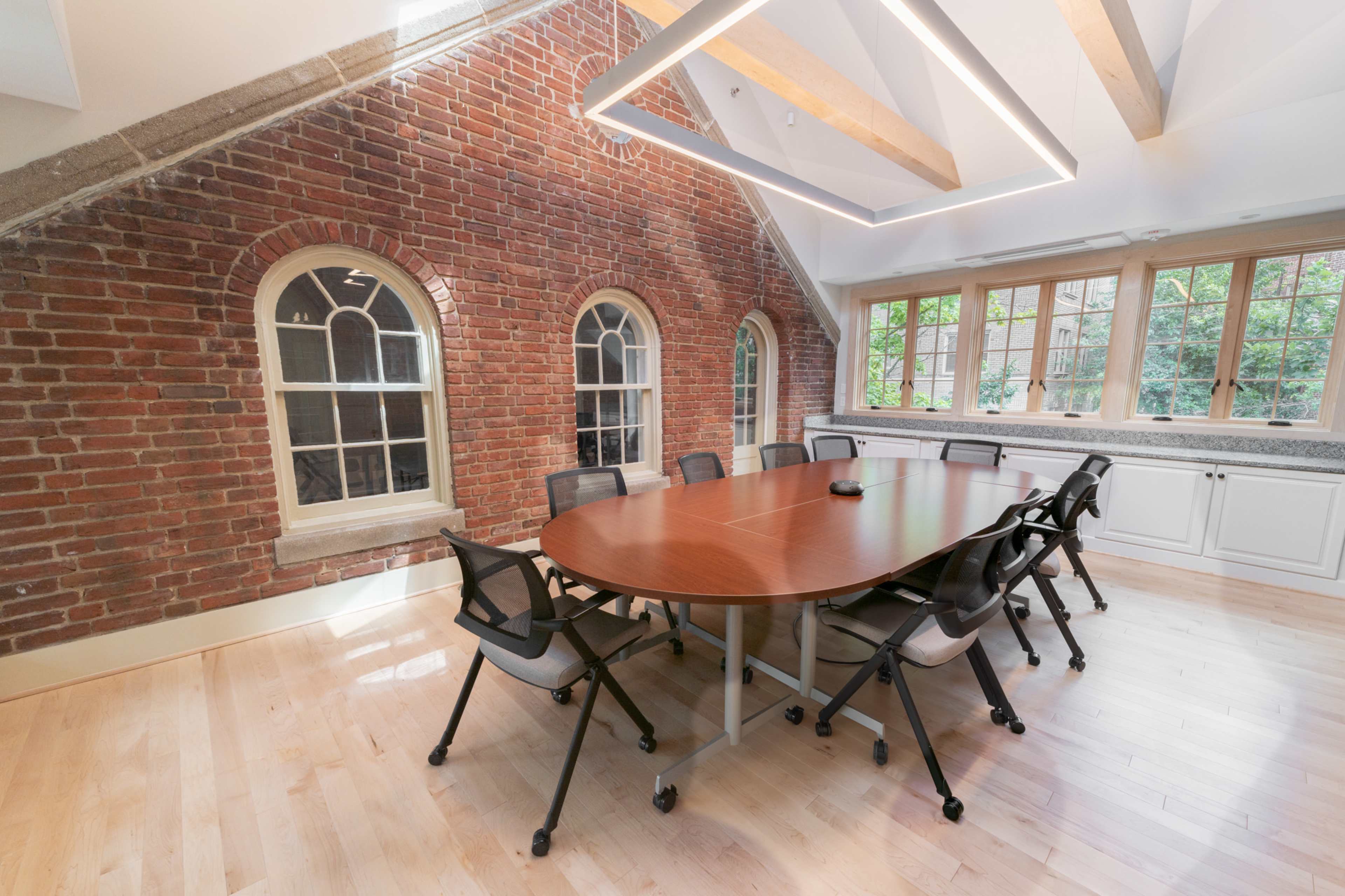 A modern conference room featuring a large wooden table surrounded by chairs, with exposed brick walls and large windows letting in natural light.
